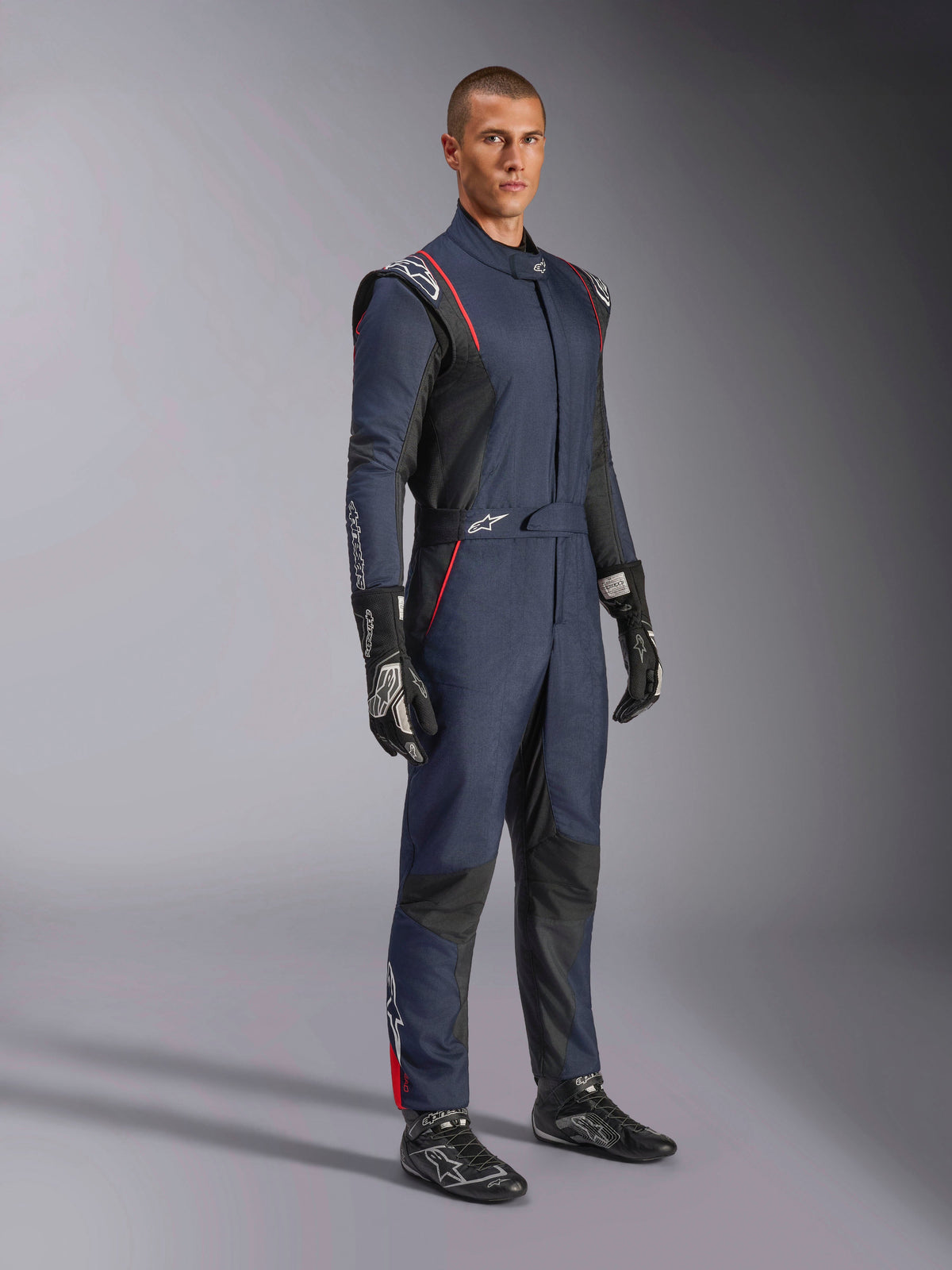 Alpinestars GP Tech V4-overall, racingoverall för bilsport, marinblå, svart och röd, bärs av en modell med svarta racinghandskar och stövlar, med svarta paneler och röda detaljer, professionell motorsportutrustning