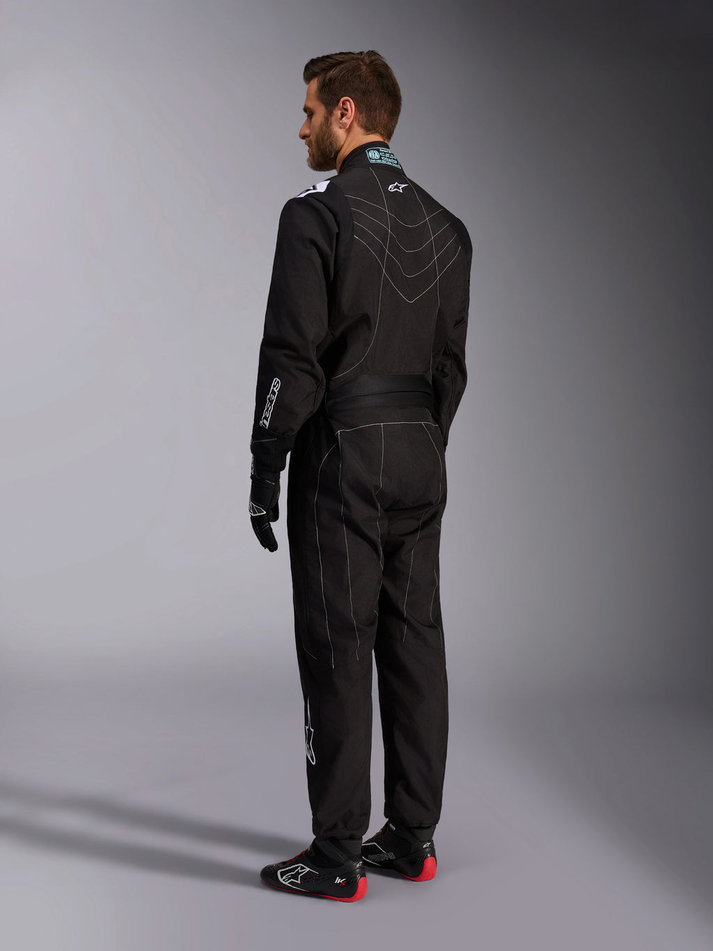 Alpinestars KMX-9 V3-dräkt, svart och vit kartingoverall, trekvartsvy bakifrån på en manlig modell, med vita kontrastsömmar, stretchpaneler på rygg och axlar, vita logotyper, bärs med svarta racinghandskar och svarta skor med röda sulor.