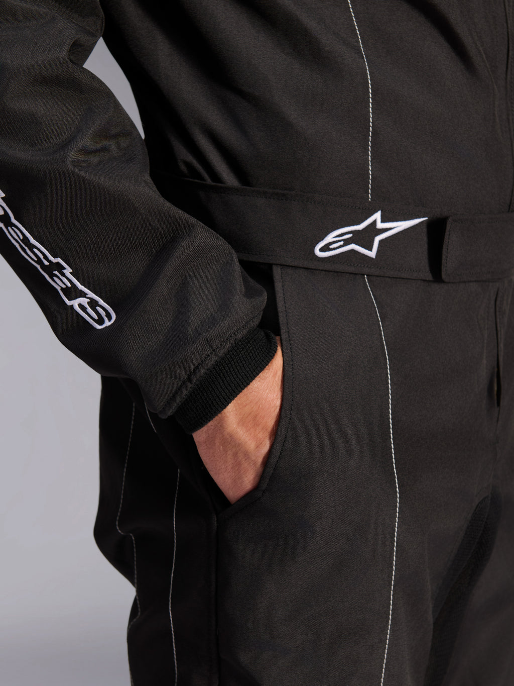 Alpinestars KMX-9 V3-dräkt, svart och vit, närbild på justerbart midjebälte med broderad logotyp, sidoficka, vita kontrastsömmar på svart tyg, teknisk detalj på kartingdräkt