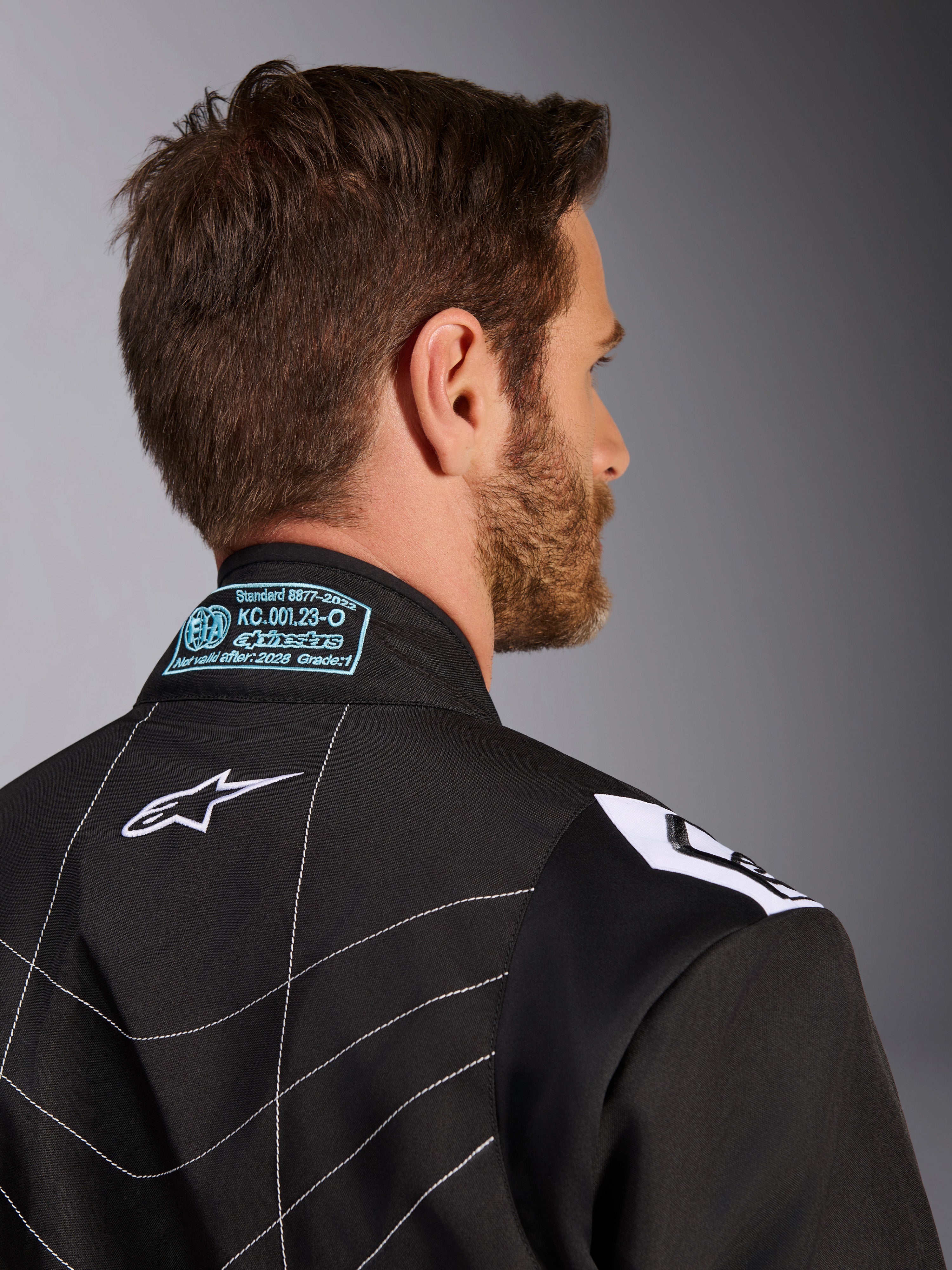 Alpinestars KMX-9 V3-dräkt, svart och vit, närbild på kragens baksida och axelpartiet, med ljusblå broderade FIA-homologeringsdetaljer, vit stjärnlogotyp och vita kontrastsömmar.