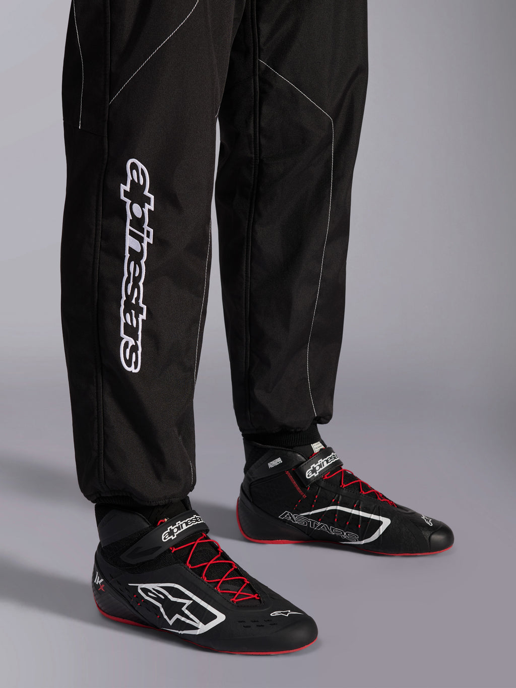 Alpinestars KMX-9 V3-dräkt, kartingdräkt, svart och vit, vy över underbenen som visar stor vit Alpinestars-logotyp, bärs med svarta och röda racingskor, professionell motorsportutrustning för karting