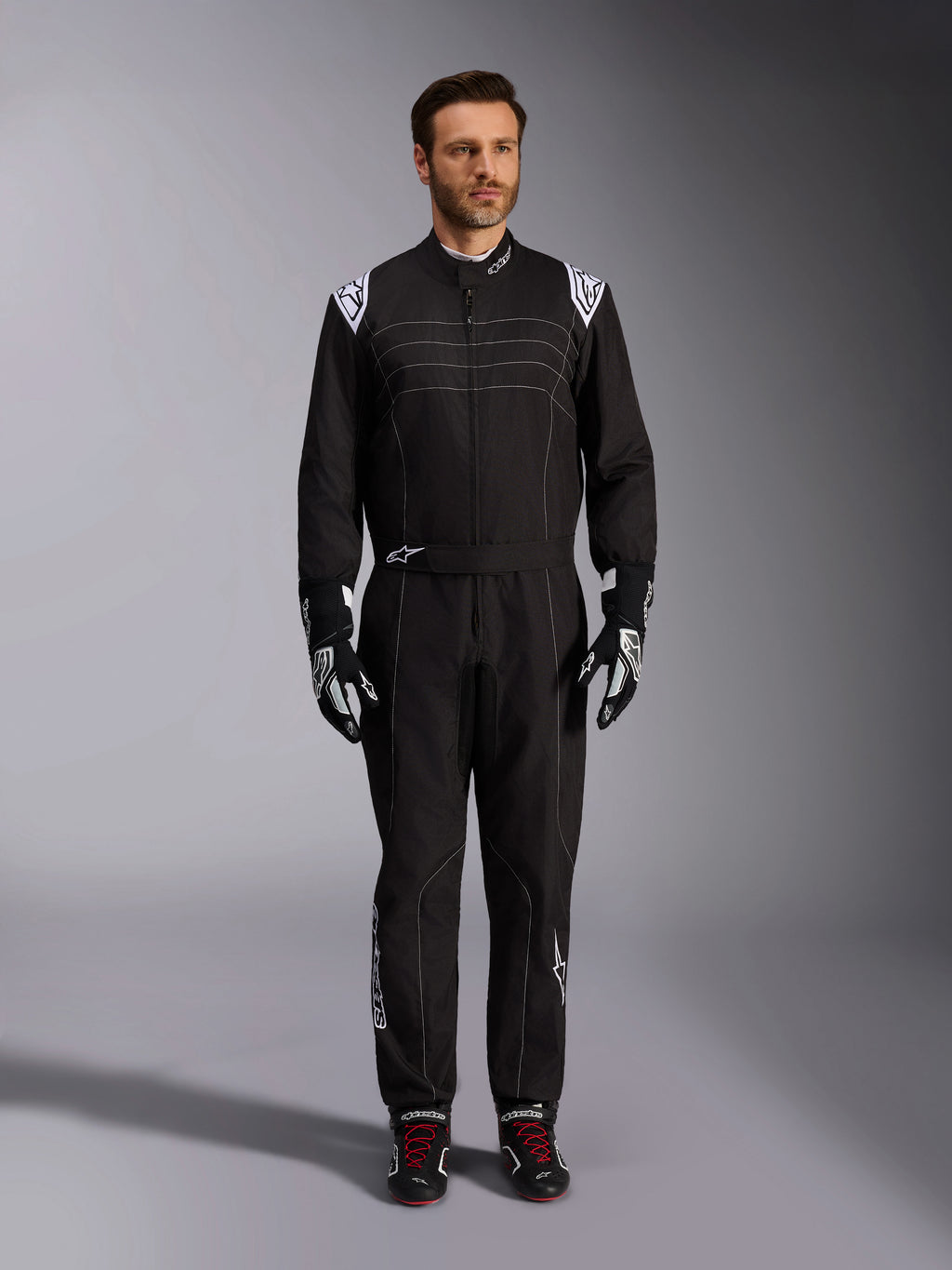 Alpinestars KMX-9 V3-ställ, svart och vit kartingoverall, helfigur på en modell, med vita axellogotyper och detaljer, stylad med svarta och vita racinghandskar och svarta skor med röda skosnören, athletic-regular passform för professionell kartingutrustning.