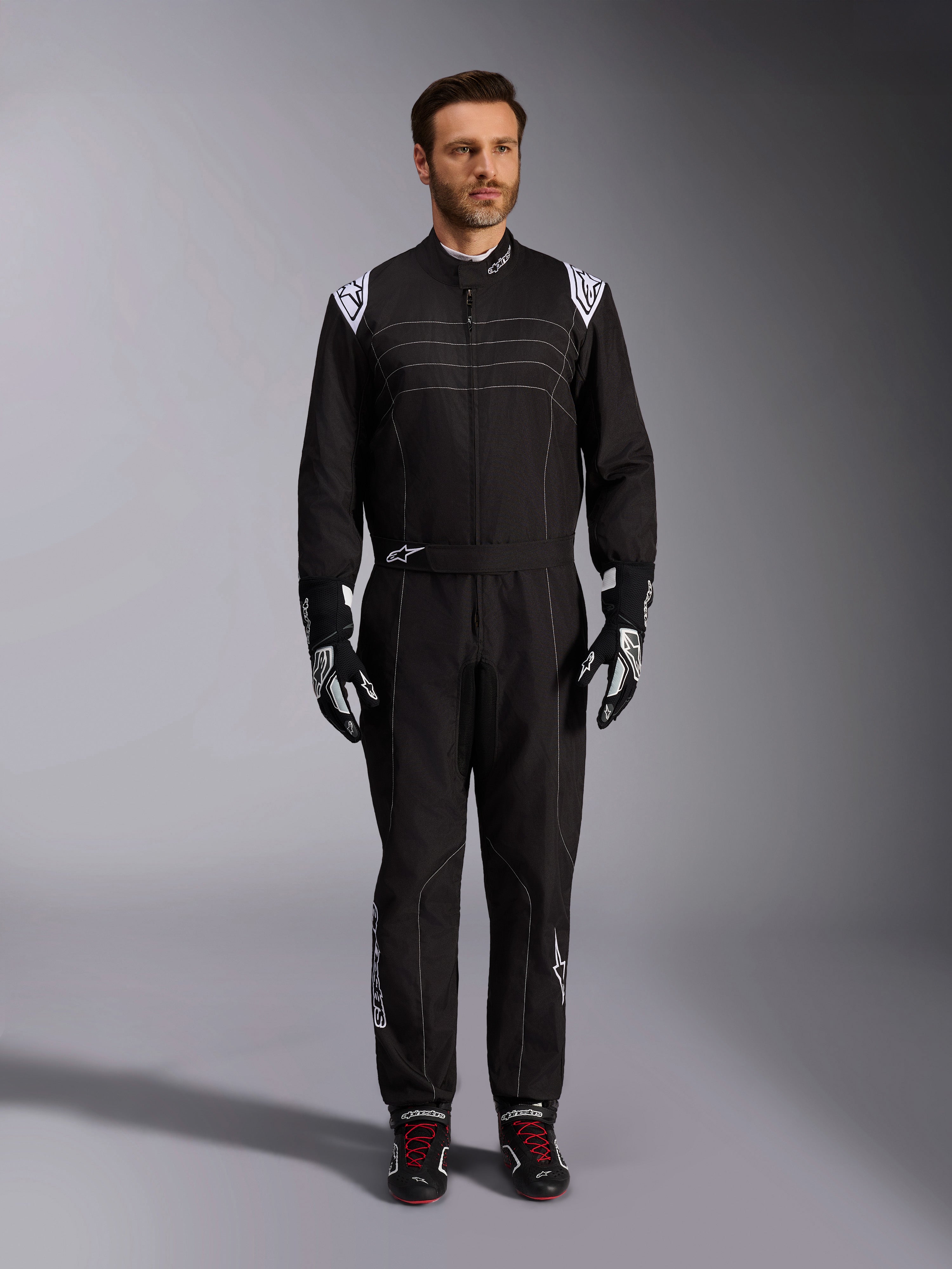 Alpinestars KMX-9 V3-ställ, svart och vit kartingoverall, helfigur på en modell, med vita axellogotyper och detaljer, stylad med svarta och vita racinghandskar och svarta skor med röda skosnören, athletic-regular passform för professionell kartingutrustning.