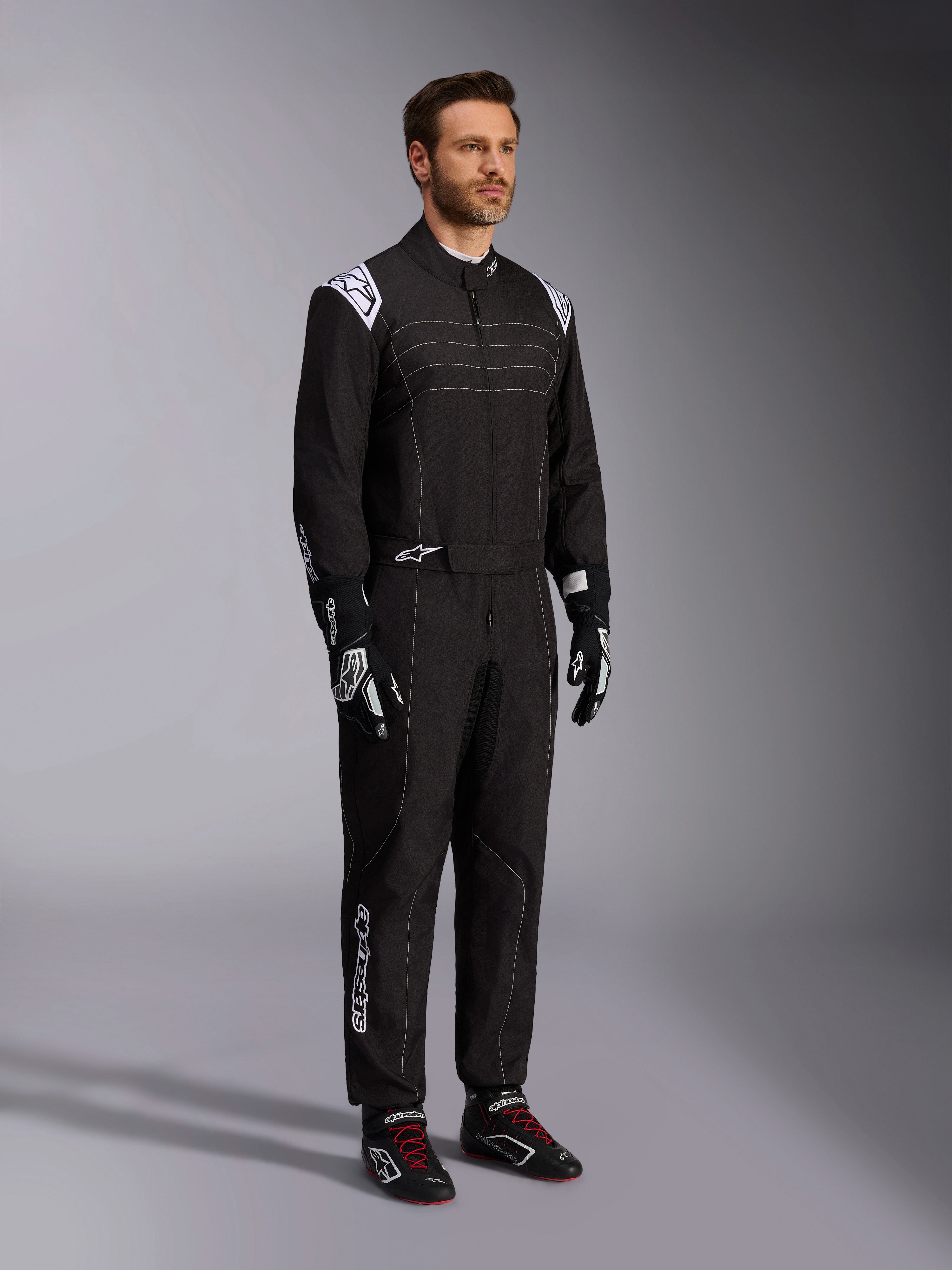 Alpinestars KMX-9 V3 kartingoverall, svart och vit, manlig modell i komplett racingoverall med vit Alpinestars-logotyp på axelklaffarna, kombinerad med svarta och vita racinghandskar och svarta kartingskor med röda skosnören, studiobild som visar fram- och sidoprofil