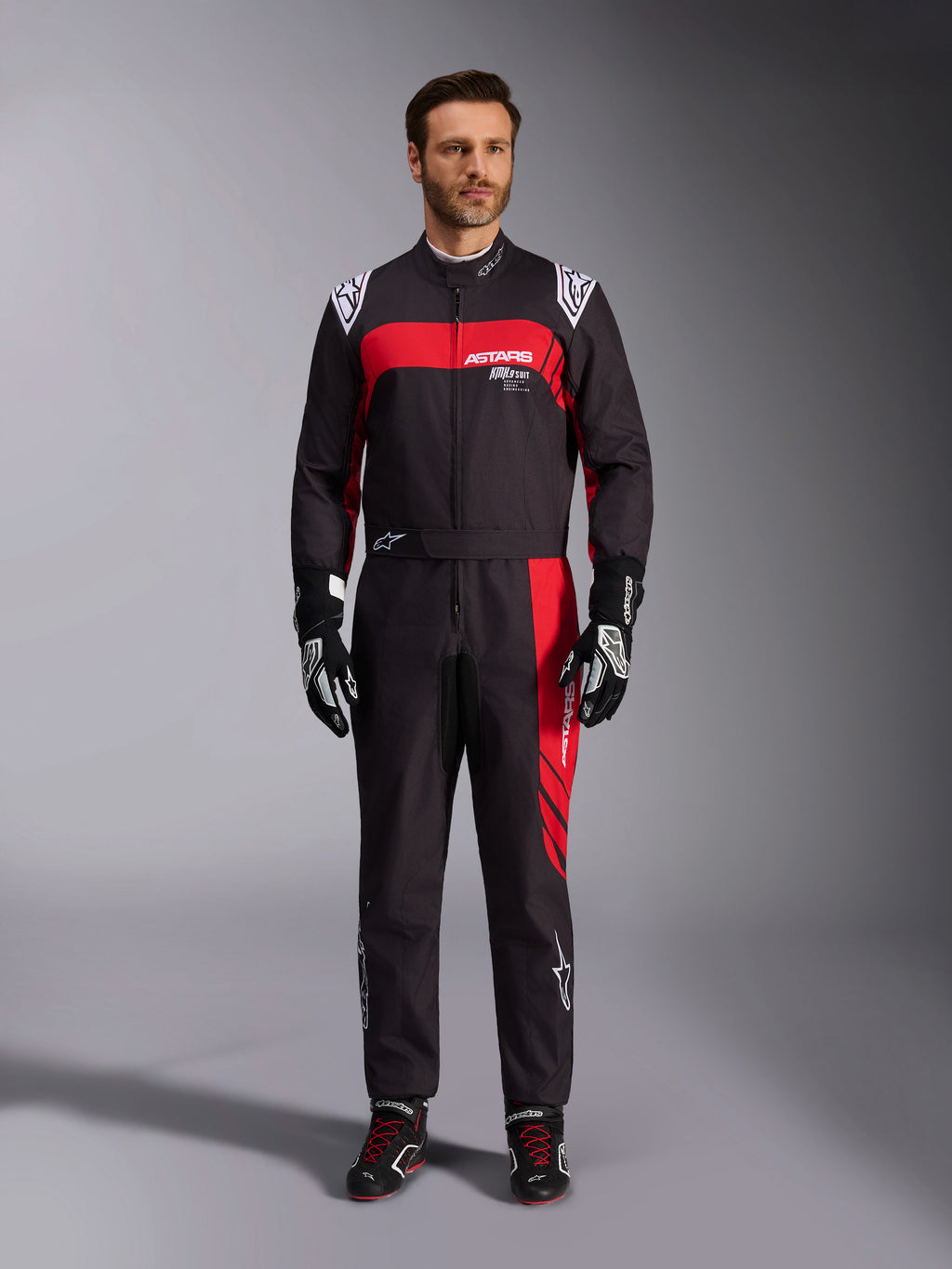 Alpinestars KMX-9 V3 Graphic 3-dräkt, kartingdräkt, svart och röd, helkroppsbild framifrån på en modell, med framträdande röda grafiska paneler och vit logotyp, kombinerad med svarta racinghandskar och skor, professionell motorsportutrustning.