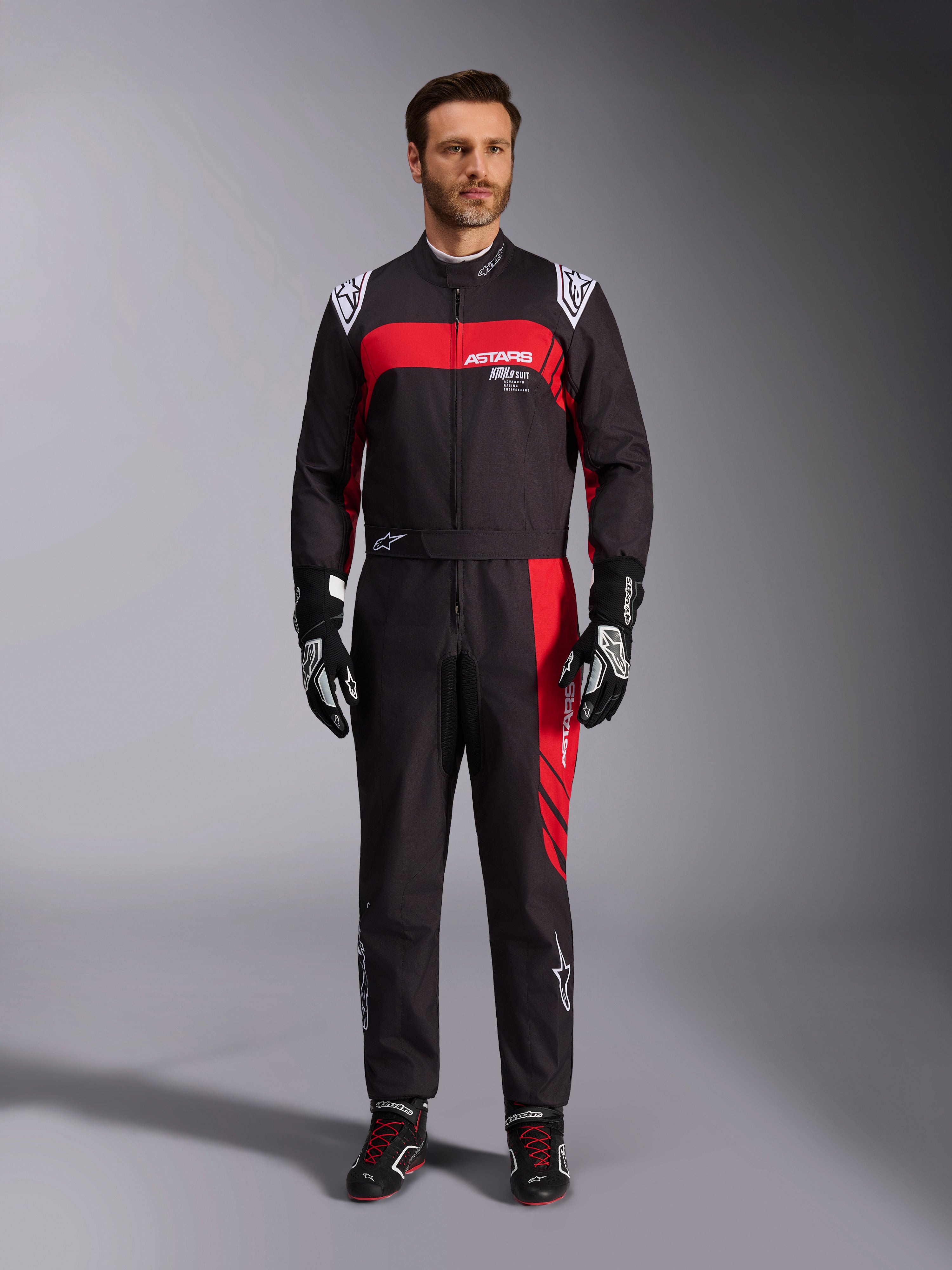 Alpinestars KMX-9 V3 Graphic 3-dräkt, kartingdräkt, svart och röd, helkroppsbild framifrån på en modell, med framträdande röda grafiska paneler och vit logotyp, kombinerad med svarta racinghandskar och skor, professionell motorsportutrustning.