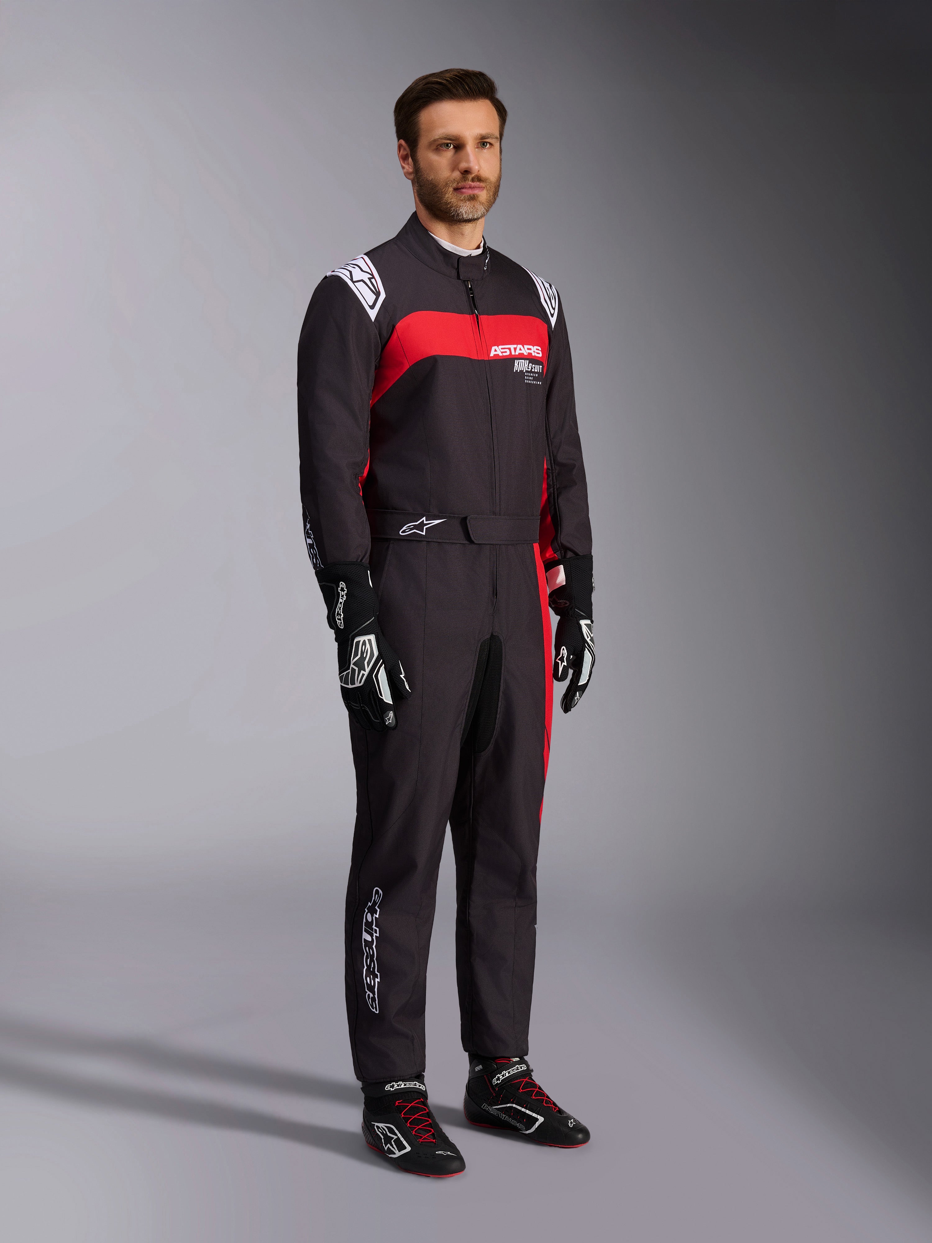 Manlig modell bär Alpinestars KMX-9 V3 Graphic 3-dräkt, kartingdräkt, svart och röd, med vita axelepåletter och röd logotyp på bröstet, kombinerat med svarta racinghandskar och skor med röda skosnören, professionell motorsportutrustning.