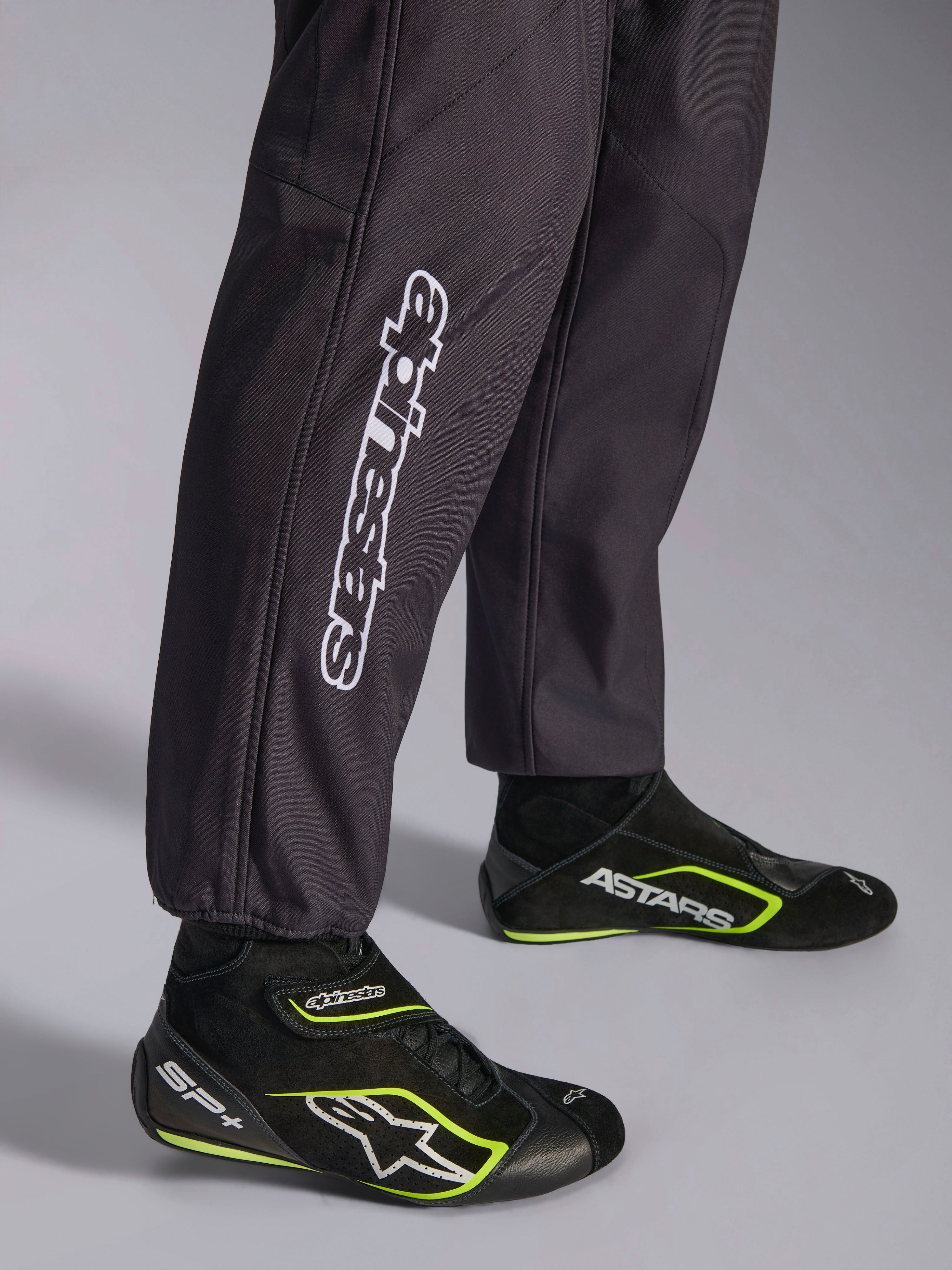 Alpinestars KMX-9 V3 Graphic 3-dräkt, kartingoverall, svart, närbild på underbenen och ankelmuddarna med vit vertikal Alpinestars-branding, kombinerad med svarta och neongula kartingskor, detalj av professionell kartingutrustning