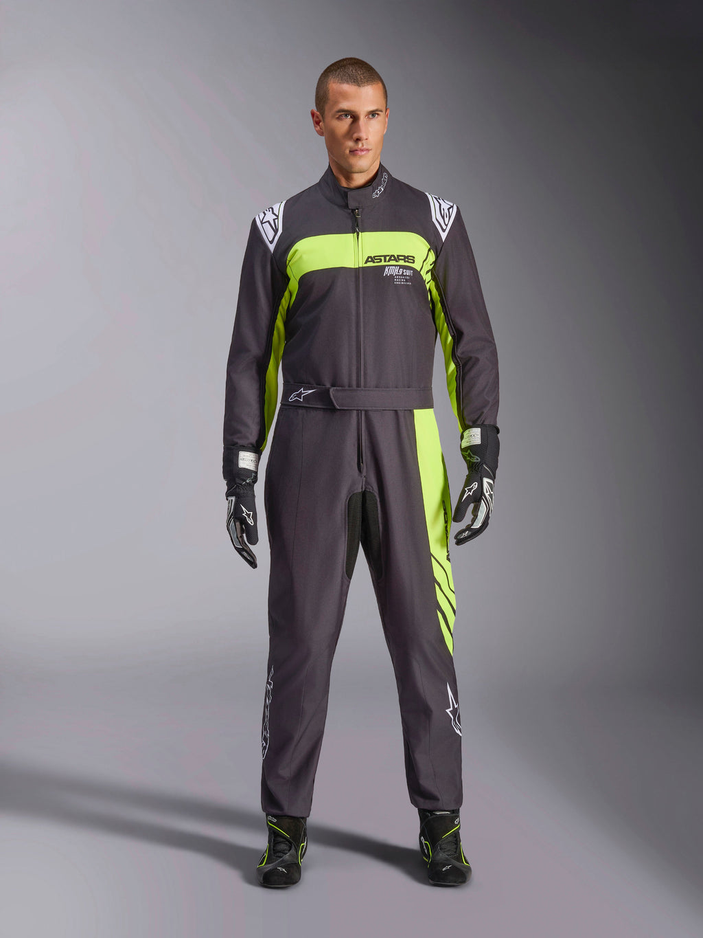 Alpinestars KMX-9 V3 Graphic 3-overall, svart och neongul, professionell kartingoverall i ett stycke som bärs av en modell, med framträdande neongula detaljer, ASTARS-logotyp på bröstet samt matchande racinghandskar och skor.