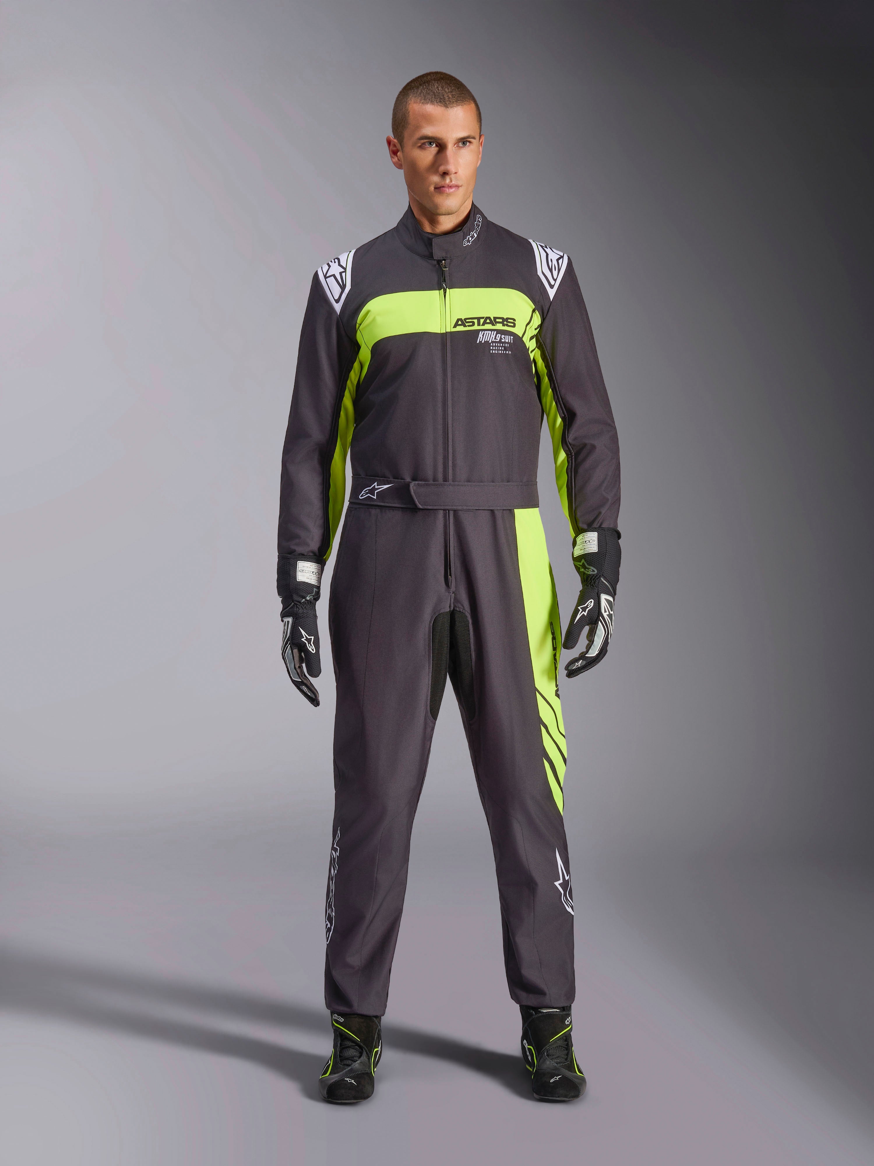 Alpinestars KMX-9 V3 Graphic 3-overall, svart och neongul, professionell kartingoverall i ett stycke som bärs av en modell, med framträdande neongula detaljer, ASTARS-logotyp på bröstet samt matchande racinghandskar och skor.