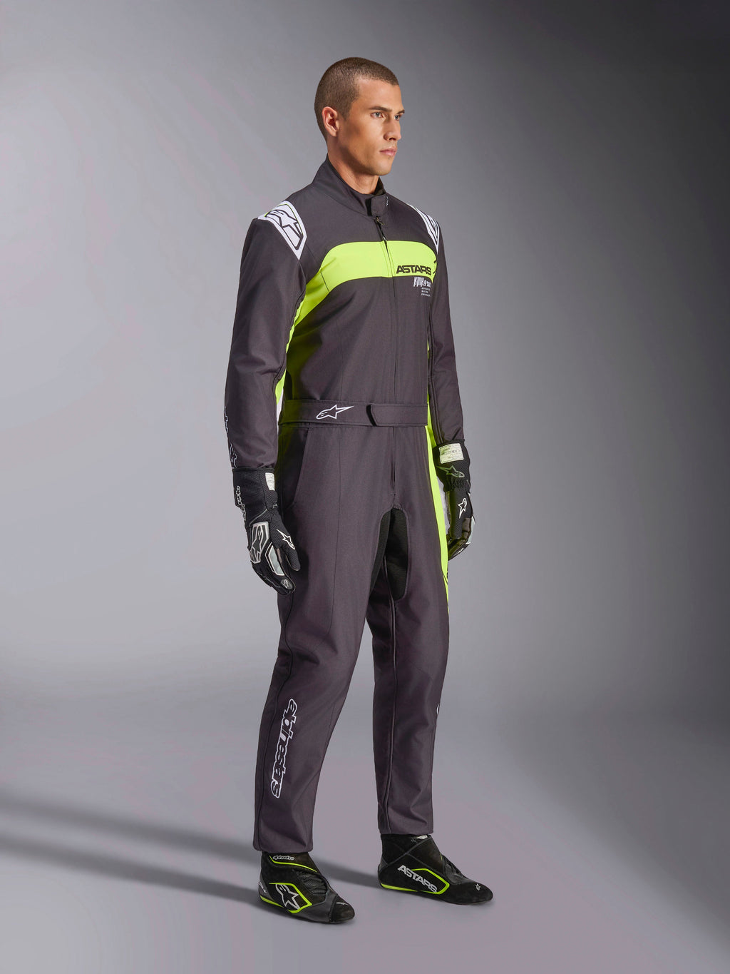 Manlig modell bär Alpinestars KMX-9 V3 Graphic 3-overall i svart och neongult, professionell kartingutrustning med livfulla neongula paneler och vita logotypdetaljer, matchad med koordinerade racinghandskar och skor, studiobakgrund