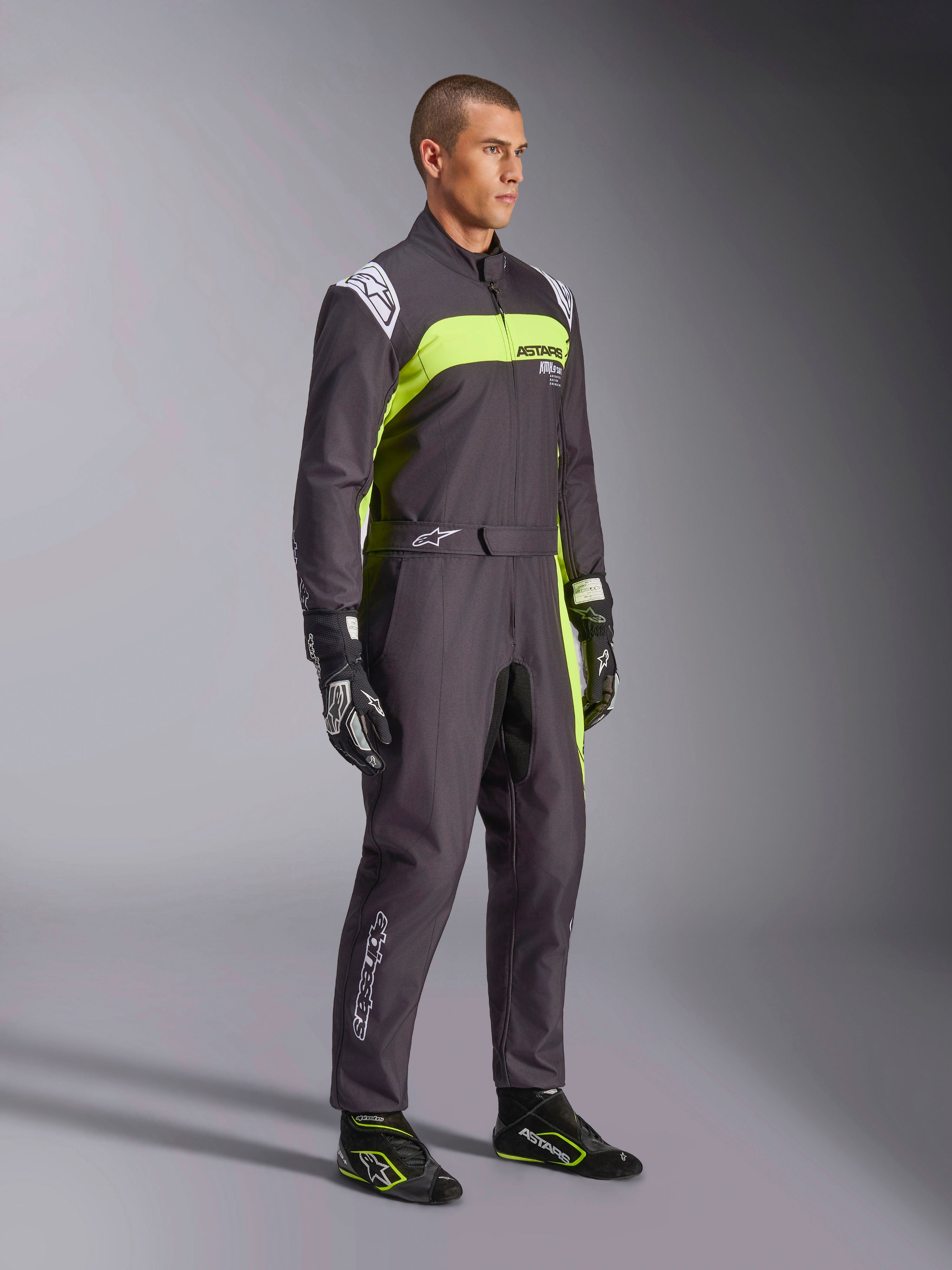 Manlig modell bär Alpinestars KMX-9 V3 Graphic 3-overall i svart och neongult, professionell kartingutrustning med livfulla neongula paneler och vita logotypdetaljer, matchad med koordinerade racinghandskar och skor, studiobakgrund