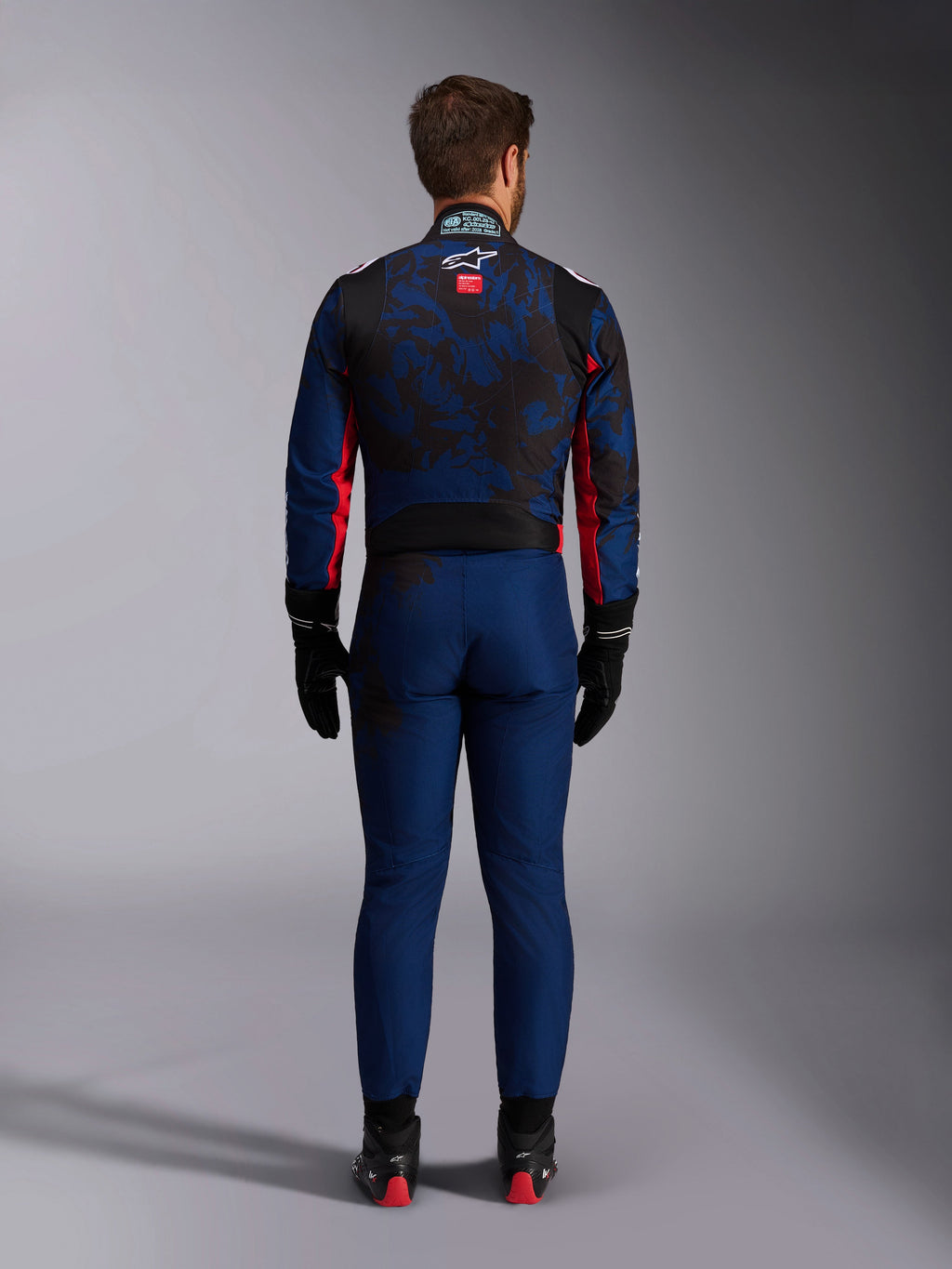 Kartingförare sedd bakifrån iförd KMX-9 V3 Graphic 5 Suit i Navy Red, med ett marinblått och svart grafiskt mönster, röda sidodetaljer och Alpinestars-logotypen på övre delen av ryggen, kombinerat med svarta racinghandskar och skor.