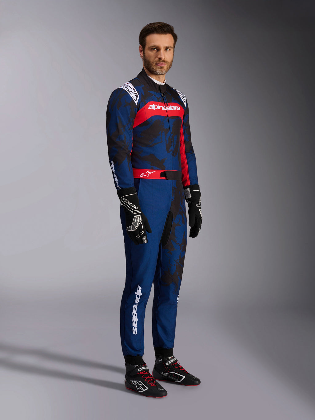 Modell som bär Alpinestars KMX-9 V3 Graphic 5 Suit i Navy Red, helkroppsbild inklusive svarta kartinghandskar och svarta racingskor, marinblå dräkt med mörk grafik i kamouflagestil och en framträdande röd bröstpanel med vit logotyp, professionell skyddsutrustning för karting
