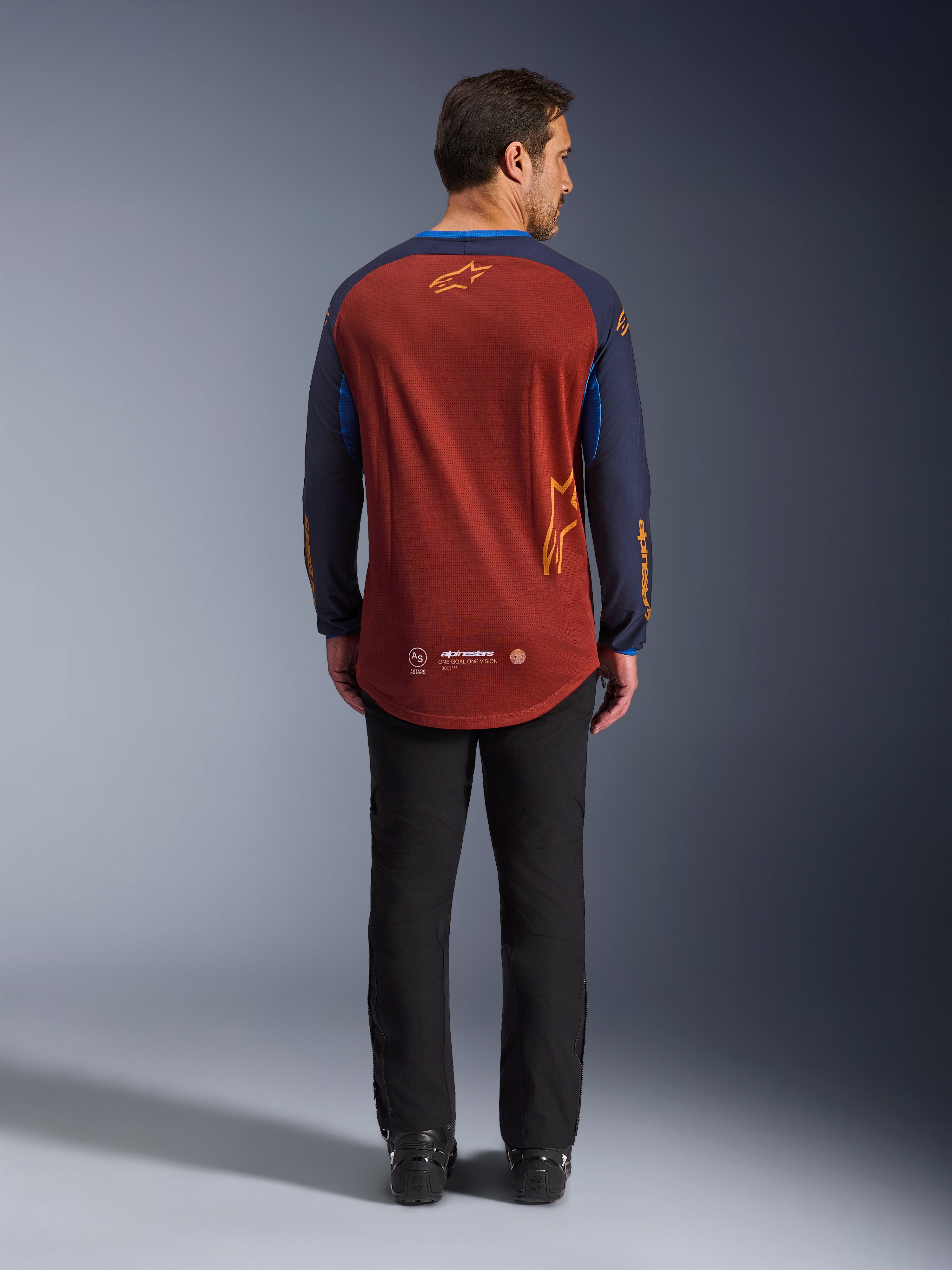 Alpinestars Maxdura Dual Jersey i Rust Brown och Mood Indigo, baksida som visar den nedsänkta bakre fållen och logotypdetaljer, bärs med svarta tekniska byxor och stövlar, designad för äventyrs- och dual-sport-motorcykelkörning.