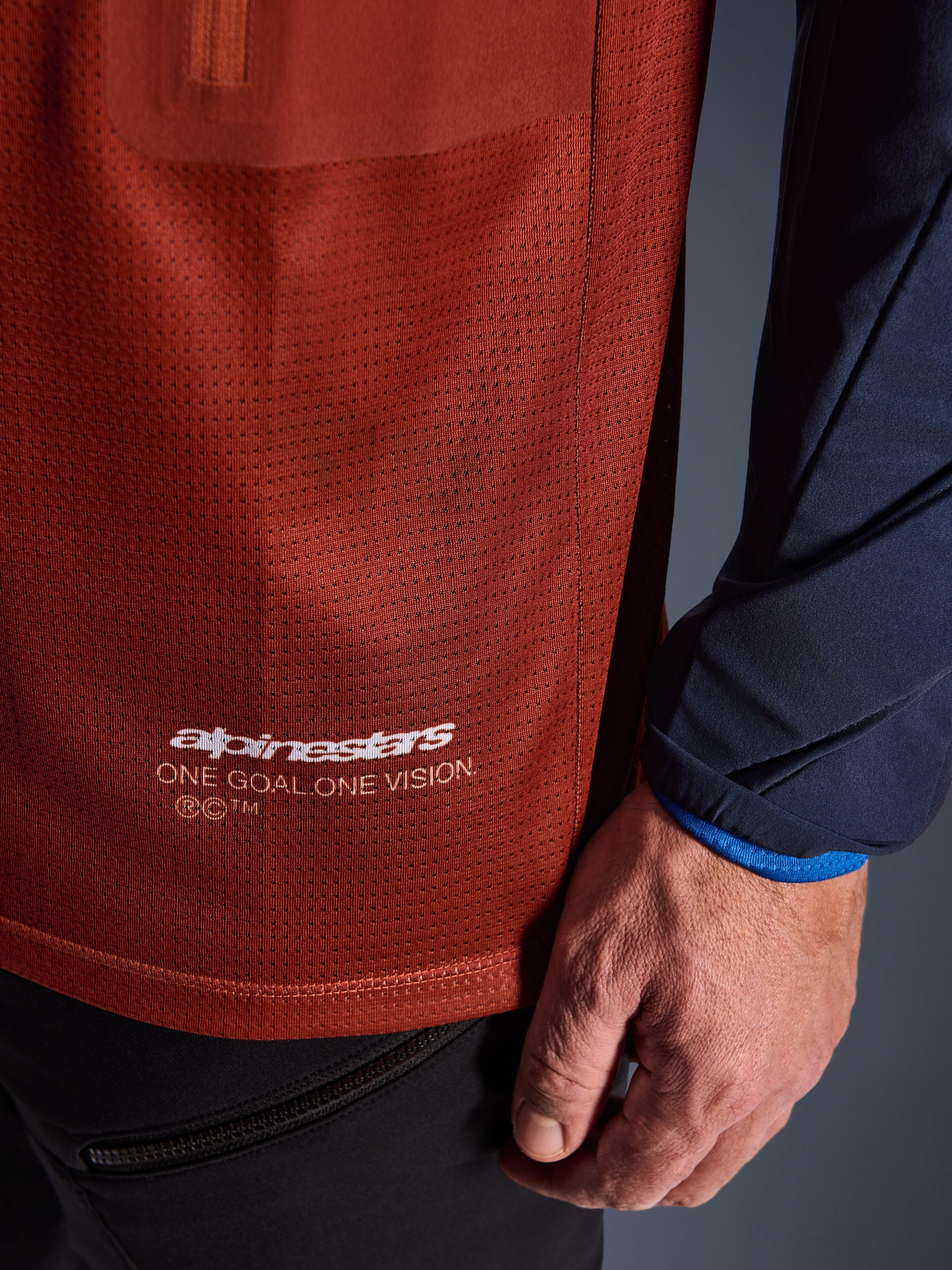 Alpinestars Maxdura Dual Jersey, äventyrströja för motorcykel, rostbrun och marinblå, närbild som visar meshtyget på torson och den marinblå vävda ärmen, vit Alpinestars 'One Goal One Vision'-logotyp på den nedre fållen.