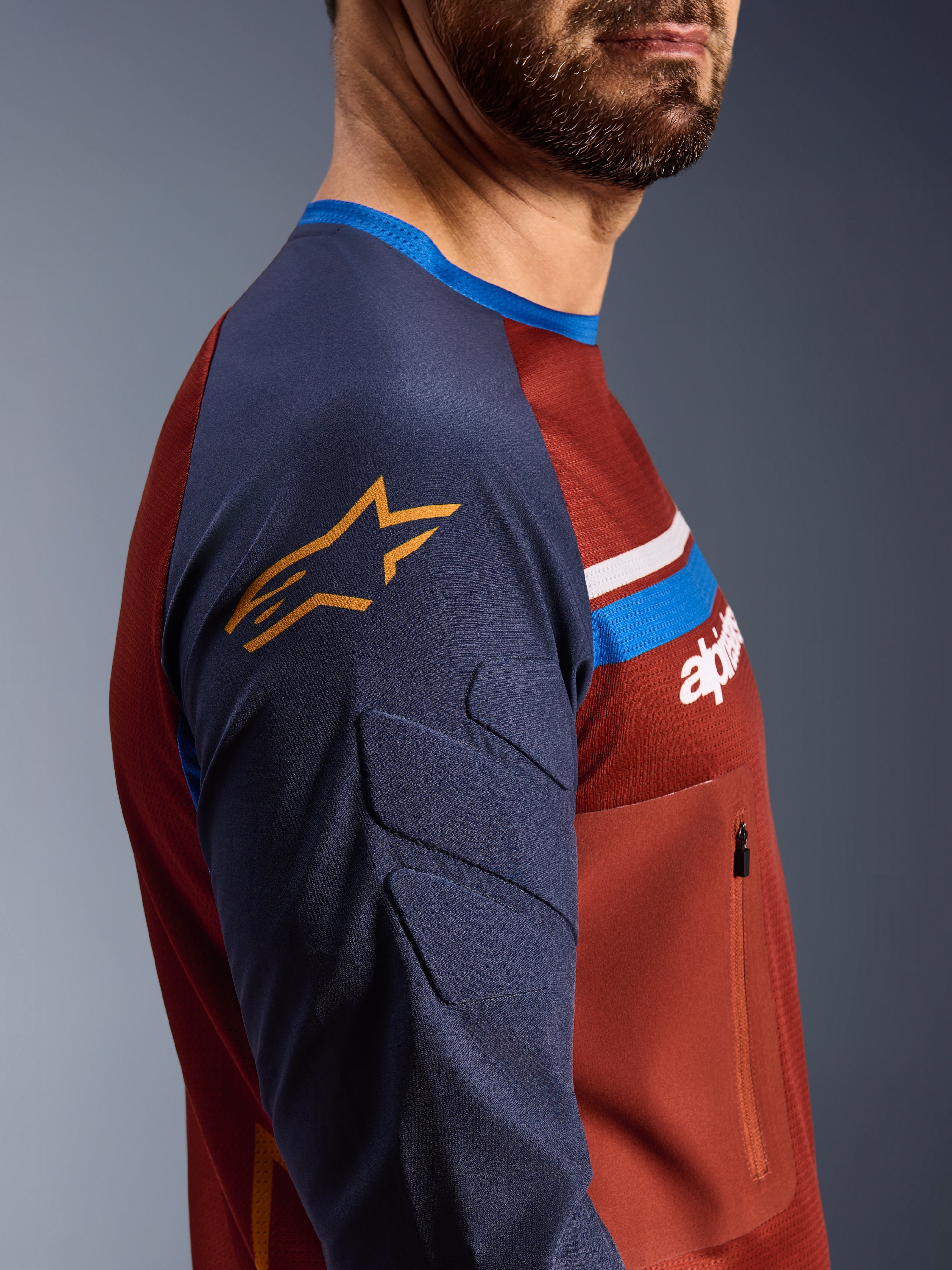 Alpinestars Maxdura Dual Jersey, rostbrun och Mood Indigo, sidoprofilvy som visar vadderade ärmar med gul stjärnlogotyp, mesh-konstruktion på torson med bröstficka med dragkedja, tekniska motorcykelkläder för adventure-körning