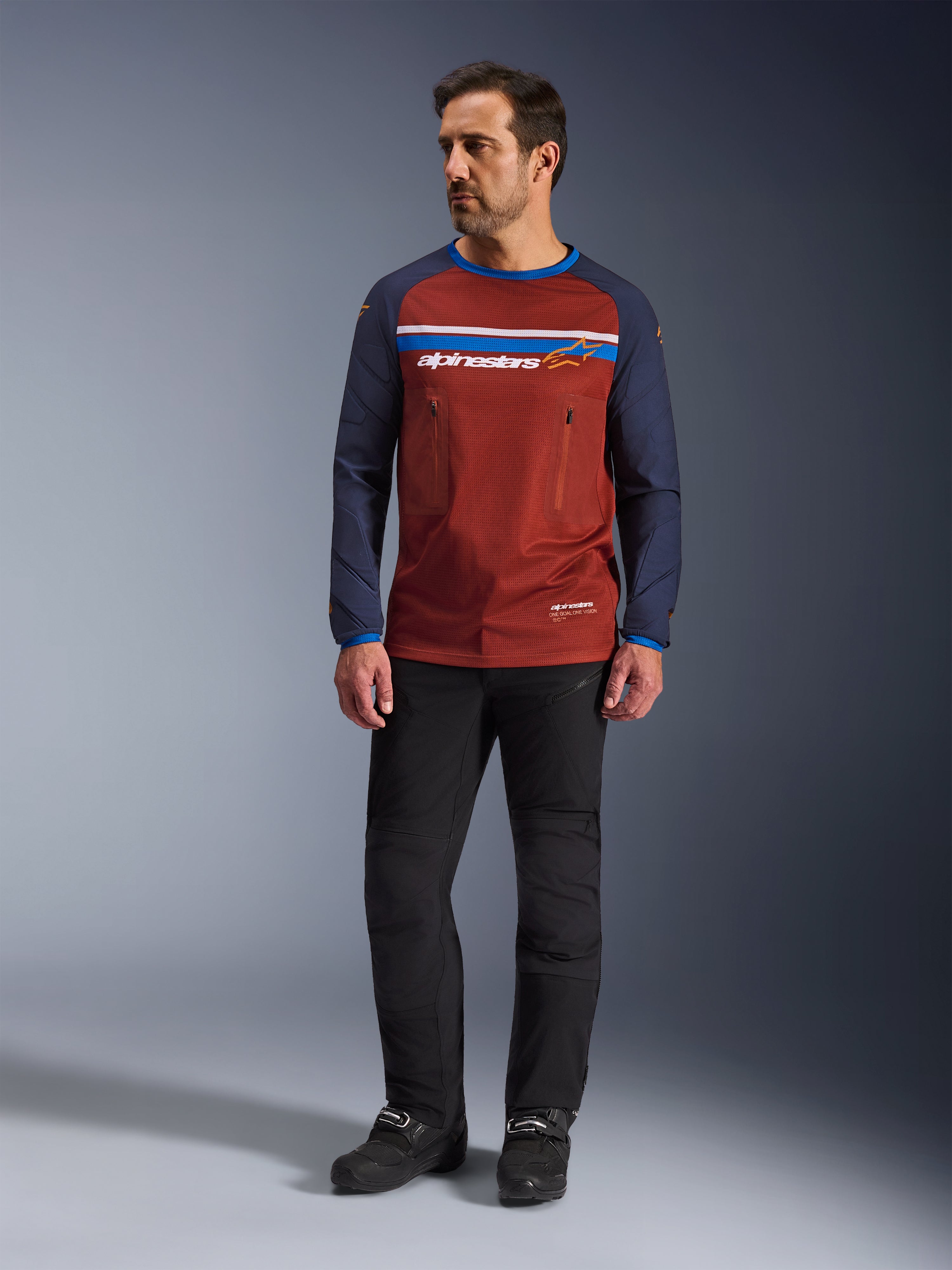 Alpinestars Maxdura Dual Jersey, rostbrun och Mood Indigo, äventyrströja för motorcykel med mesh-torso och marinblå ärmar, bärs av en modell med svarta körbyxor och svarta stövlar, teknisk dual-sport-utrustning för terrängkörning