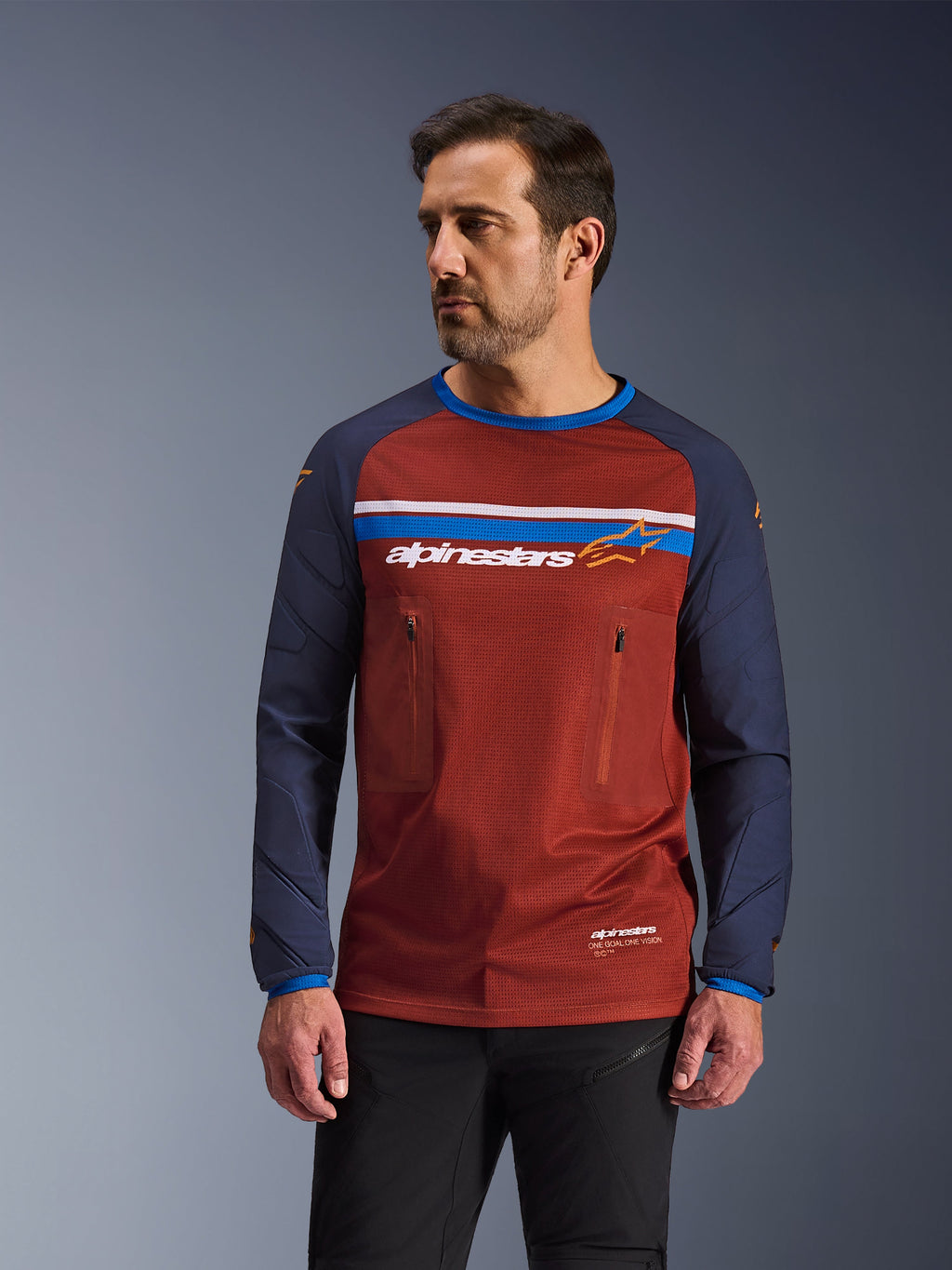 Manlig modell iklädd Alpinestars Maxdura Dual Jersey, rostbrun och marinblå äventyrsmotorcykeltröja, med dubbla bröstfickor med dragkedja, horisontell bröstrand med logotyp, teknisk mesh-torso och hållbara vävda ärmar.