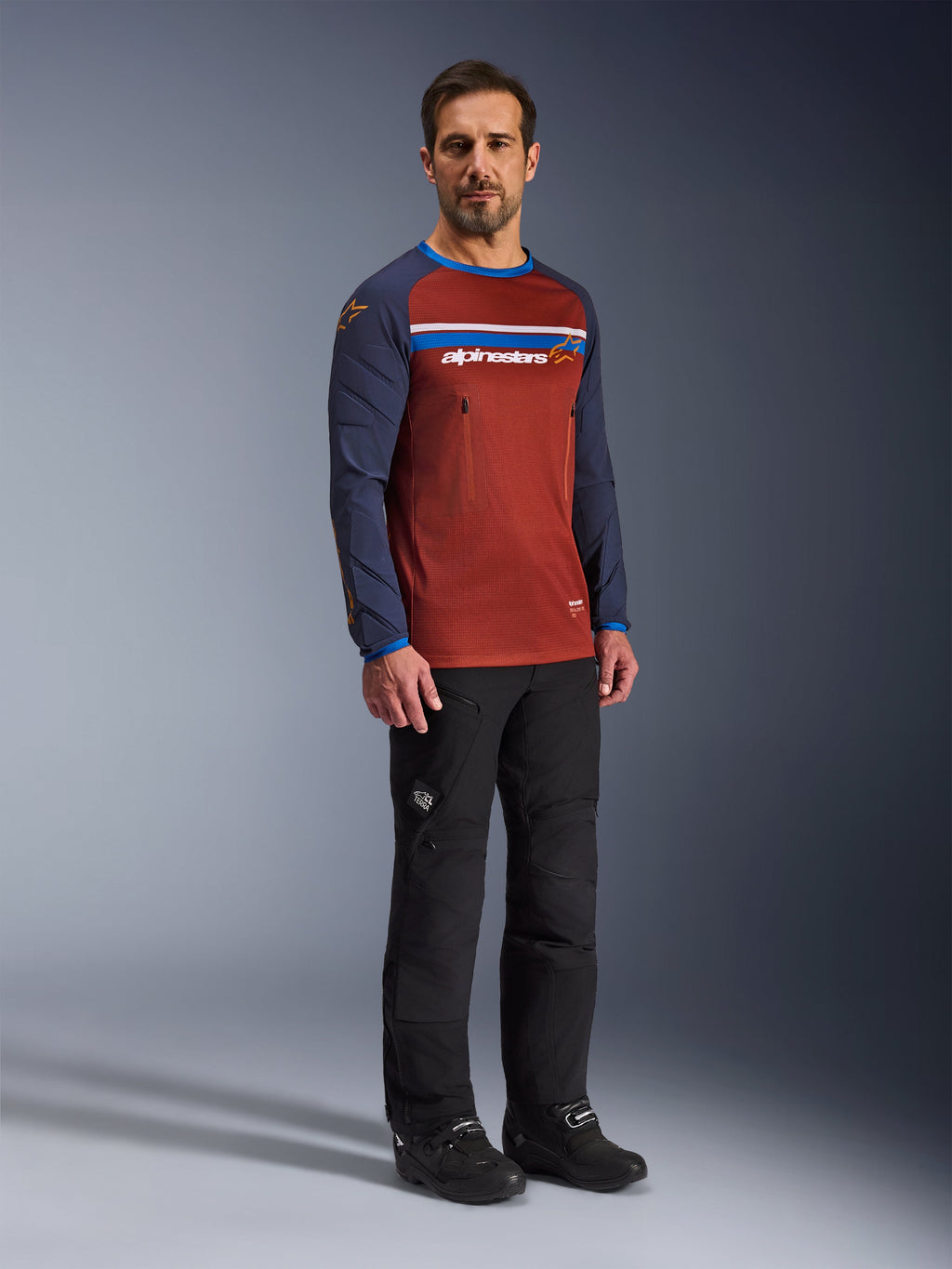 Man som bär Alpinestars Maxdura Dual Jersey i Rust Brown och Mood Indigo, med torso i mesh och vadderade ärmar, kombinerat med svarta mc-byxor och stövlar, äventyrskläder för motorcykel.