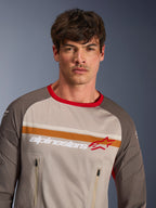 Manlig modell i Alpinestars Maxdura Dual Jersey i färgkombinationen Aluminum Walnut, adventure-motorcykeltröja med helmasktorso, nötningsresistenta ärmar, röd krage och bröstfickor med dragkedja, orangea och vita ränder på bröstet med Alpinestars-logotyp.