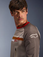 Manlig modell som bär Alpinestars Maxdura Dual Jersey i Aluminum Walnut, med ventilerad mesh-bål, röd krage och orange bröstrand. Närbild på nötningsbeständig ärm med integrerad vaddering och bröstficka med dragkedja för äventyrskörning.