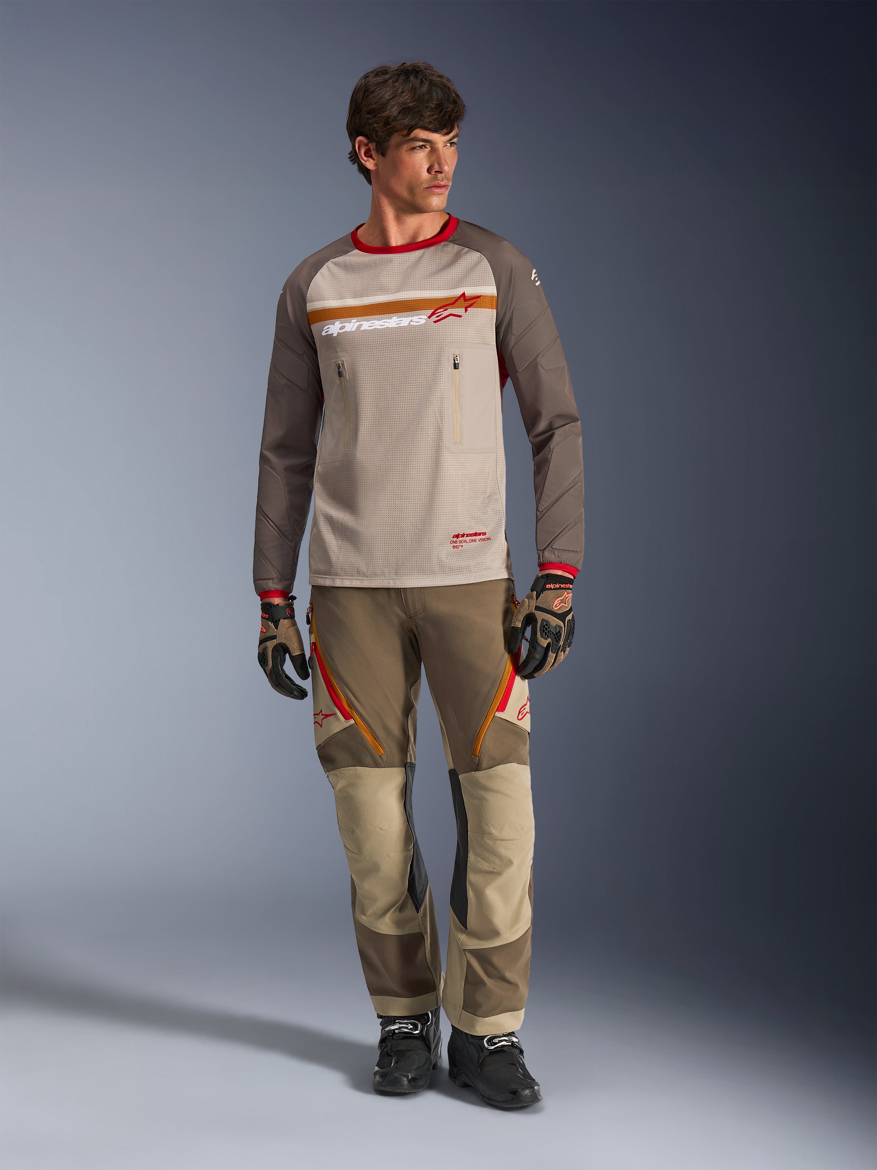 Alpinestars Maxdura Dual Jersey i Aluminum Walnut, adventure-motorcykeltröja med beige mesh-torso och bruna ärmar, röd krage och vågrätt bröstband med Alpinestars-logotyp, buren med matchande adventure-byxor och handskar