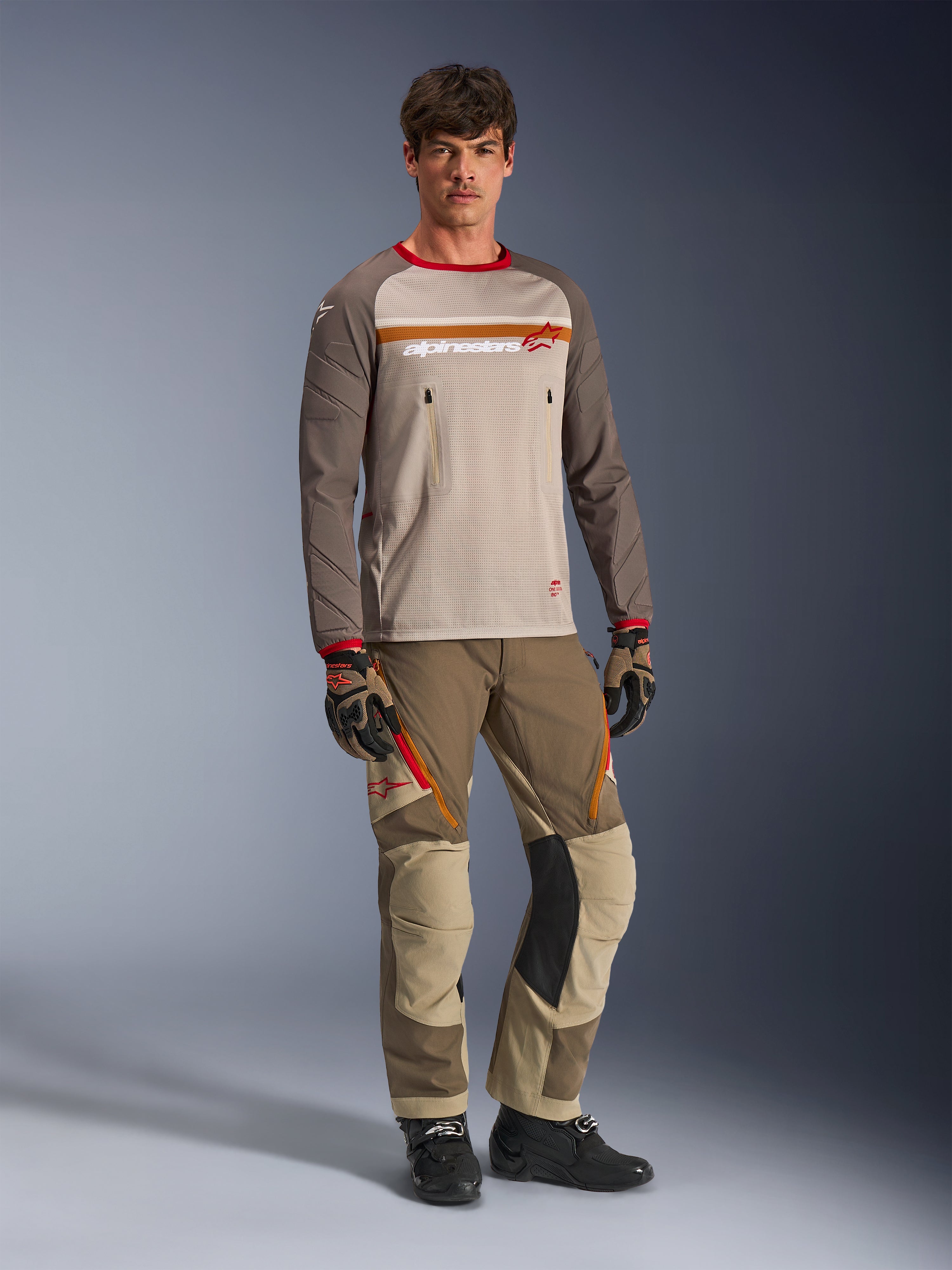 Modell som bär Alpinestars Maxdura Dual Jersey i Aluminum Walnut, med grå mesh-torso, bruna ärmar med röda muddar och bröstfickor med dragkedja, stylad med matchande off-road-utrustning för adventure-körning.