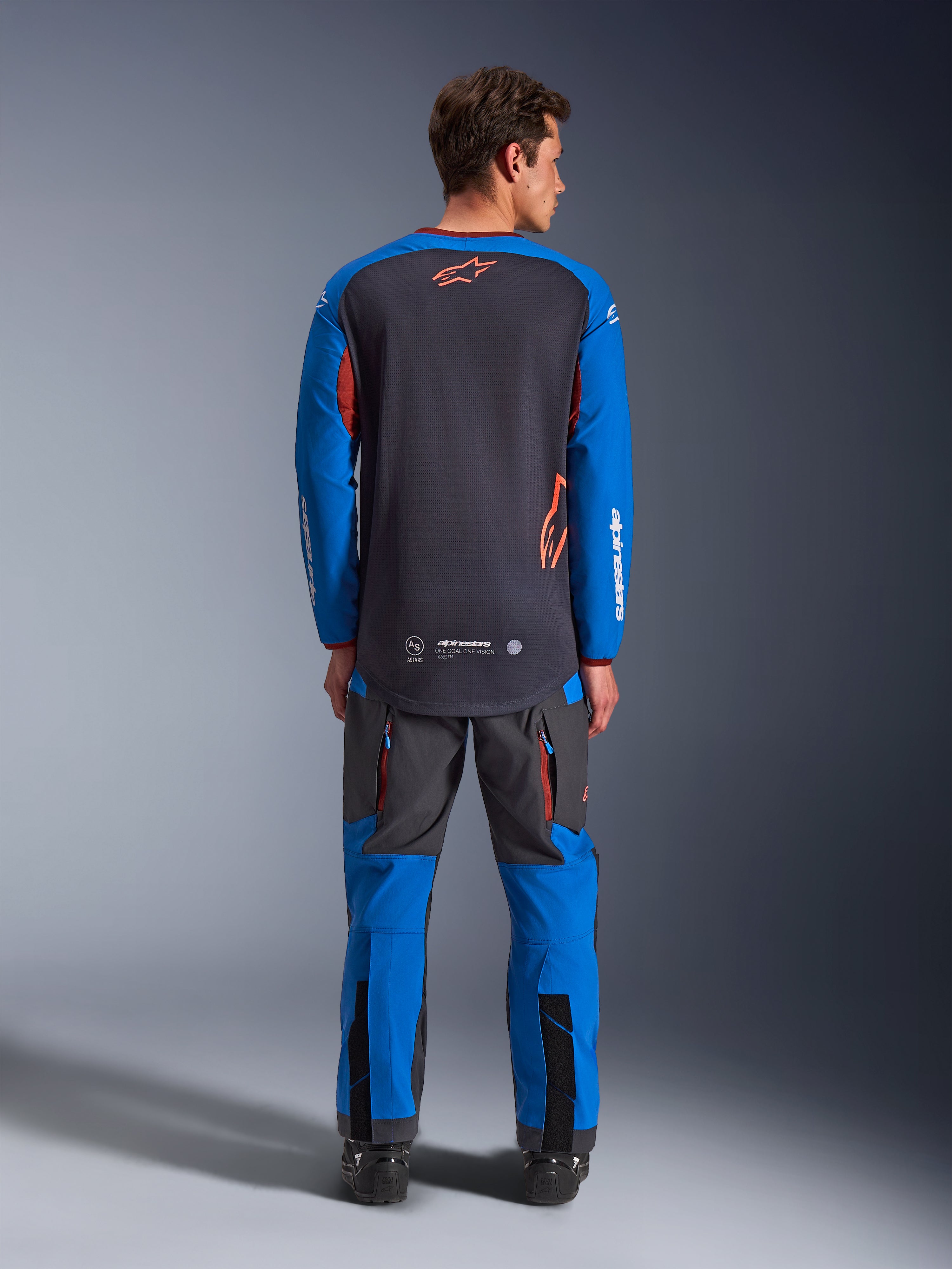 Alpinestars Maxdura Dual Jersey i mörkgrått och royalblått, ryggvy som visar bål i helmask och orange stjärnlogotyp, royalblå ärmar med vit logotyp, bärs tillsammans med matchande grå och blå adventure-byxor, högpresterande utrustning för dual-sport-körning