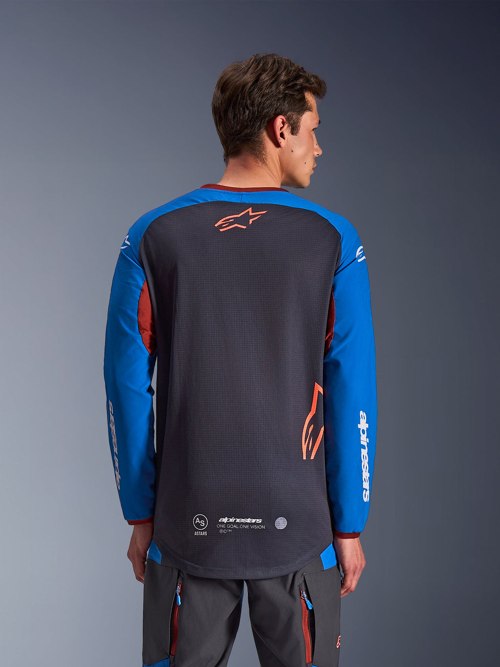 Alpinestars Maxdura Dual Jersey i Deep Gray och Royal Blue, baksida på manlig modell, mesh-konstruktion på bålen med orange Astars-logotyp, långa ärmar med vit logotyp, vinröd krage och muddar, bärs med grå tekniska byxor, motorcykelutrustning för dual-sport och äventyr