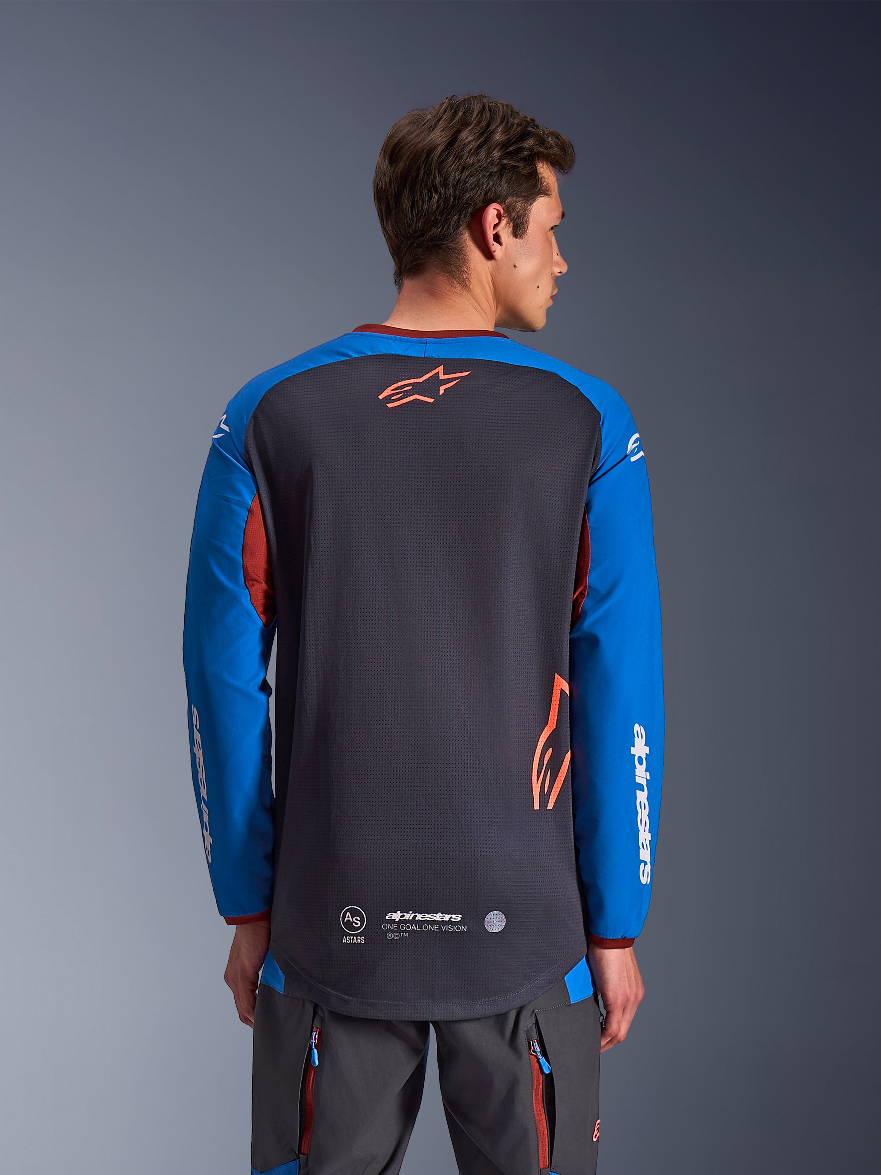 Alpinestars Maxdura Dual Jersey i Deep Gray och Royal Blue, baksida på manlig modell, mesh-konstruktion på bålen med orange Astars-logotyp, långa ärmar med vit logotyp, vinröd krage och muddar, bärs med grå tekniska byxor, motorcykelutrustning för dual-sport och äventyr