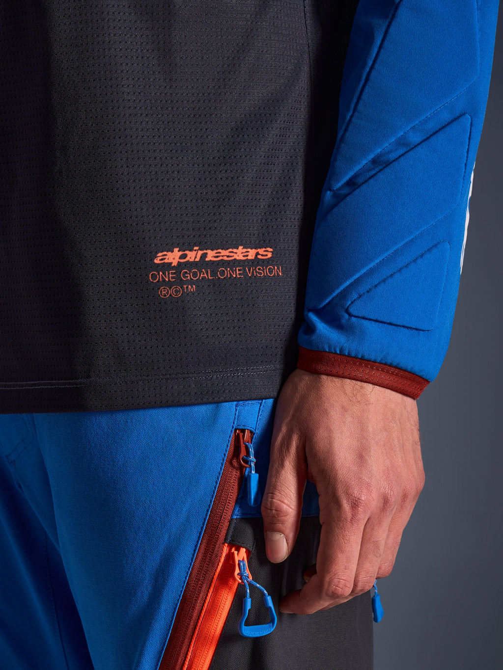 Närbild av Alpinestars Maxdura Dual Jersey i mörkgrått och kungsblått, med en orange logotyp och texten 'One Goal One Vision' på torson i mesh, vadderad ärm med förstärkta geometriska paneler, teknisk detalj för äventyrskörning