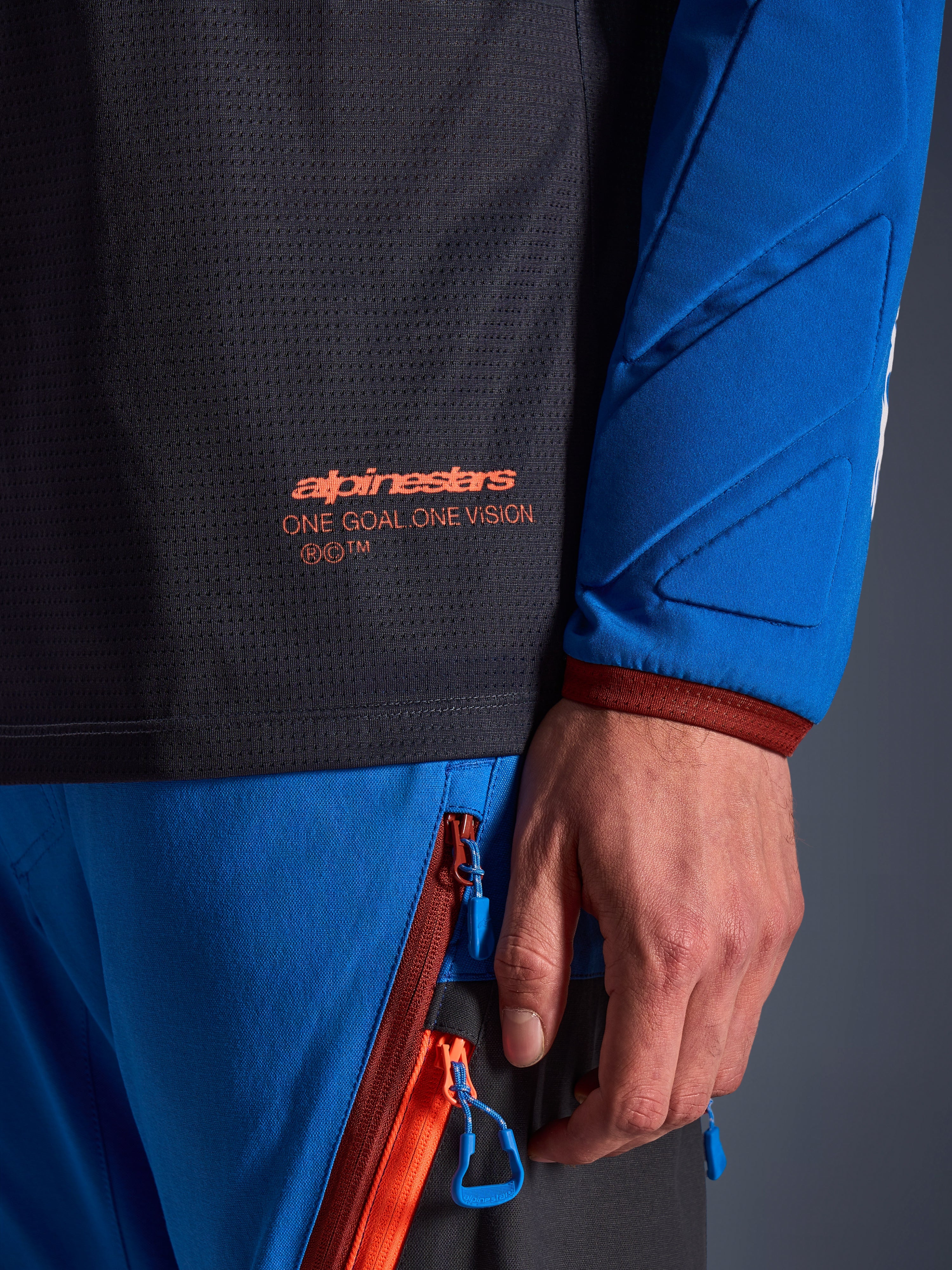 Närbild av Alpinestars Maxdura Dual Jersey i mörkgrått och kungsblått, med en orange logotyp och texten 'One Goal One Vision' på torson i mesh, vadderad ärm med förstärkta geometriska paneler, teknisk detalj för äventyrskörning