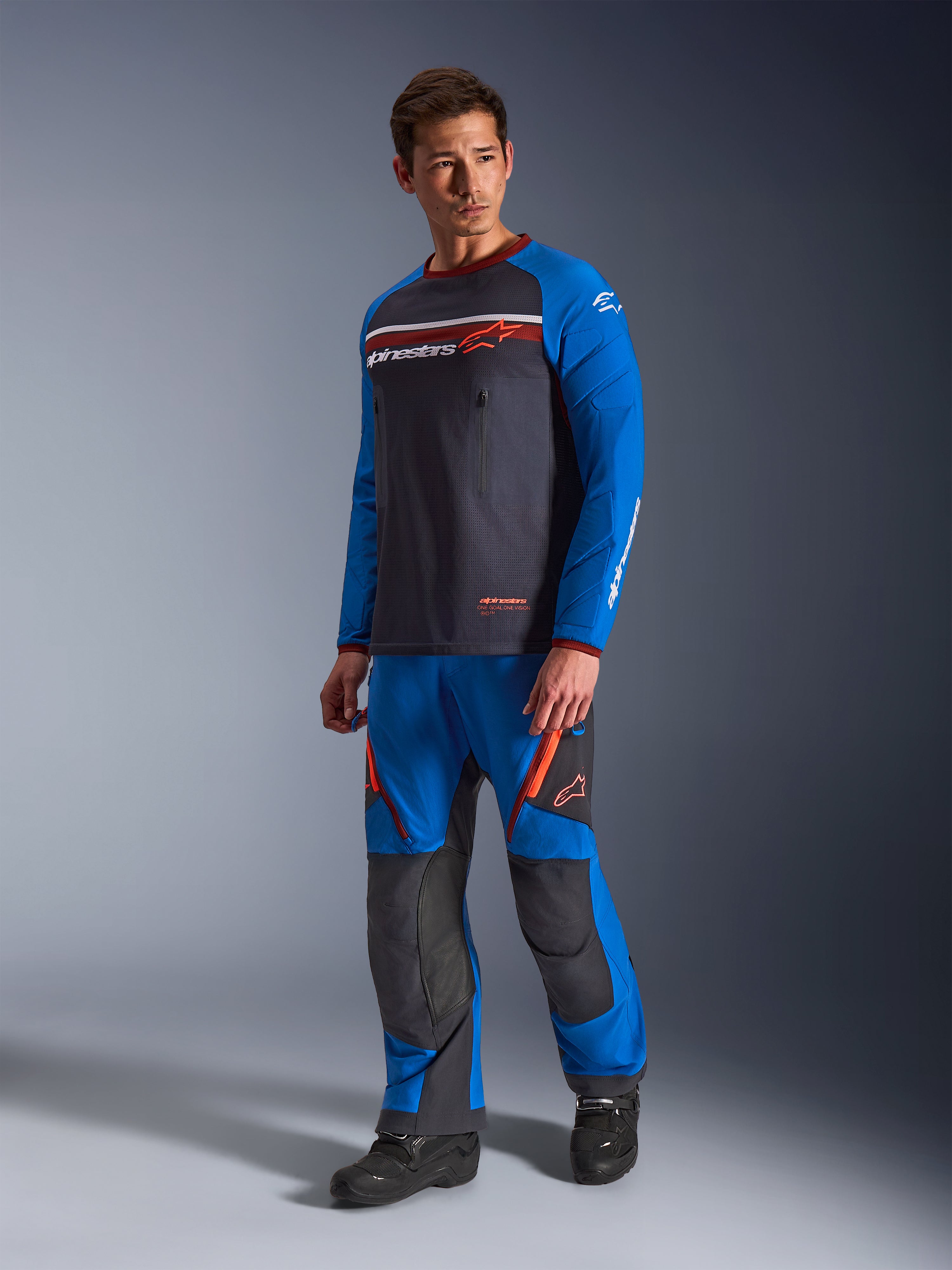 Alpinestars Maxdura Dual Jersey, mörkgrå och kungsblå, äventyrsmotorcykeltröja med mesh-bål och kungsblå ärmar, bärs av en modell med matchande off-road-körbyxor, bröstfickor med dragkedja och Alpinestars-branding