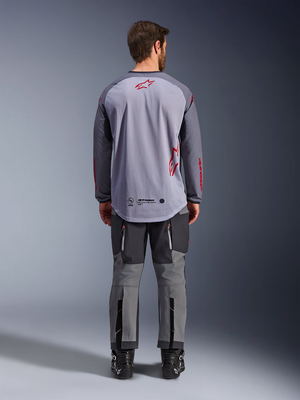 Manlig modell sedd bakifrån bär Alpinestars Maxdura Dual Jersey, mellangrå och mörkgrå äventyrströja med röda logotypdetaljer, mesh-konstruktion på ryggen, bärs med matchande grå off-road-byxor och svarta stövlar