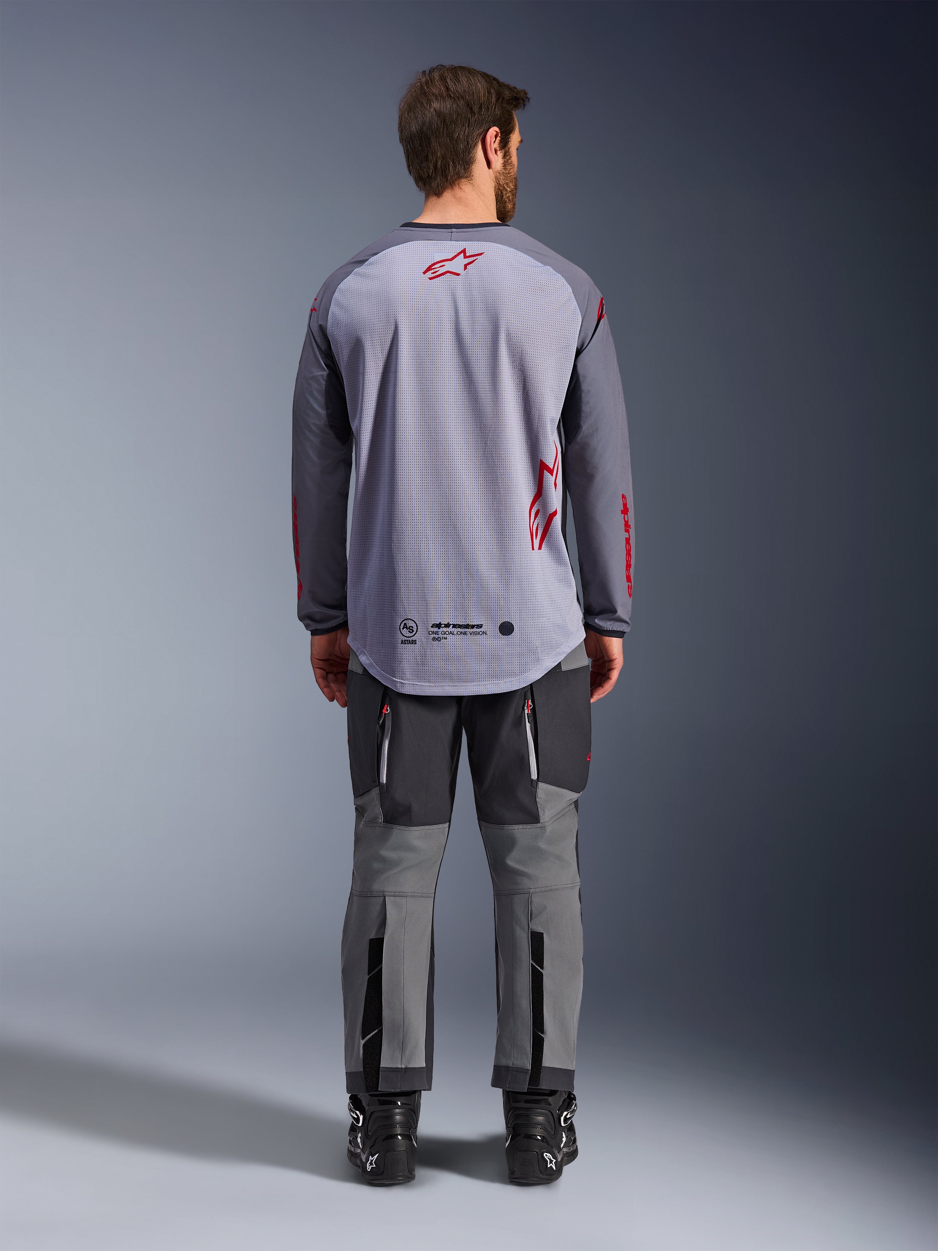 Manlig modell sedd bakifrån bär Alpinestars Maxdura Dual Jersey, mellangrå och mörkgrå äventyrströja med röda logotypdetaljer, mesh-konstruktion på ryggen, bärs med matchande grå off-road-byxor och svarta stövlar