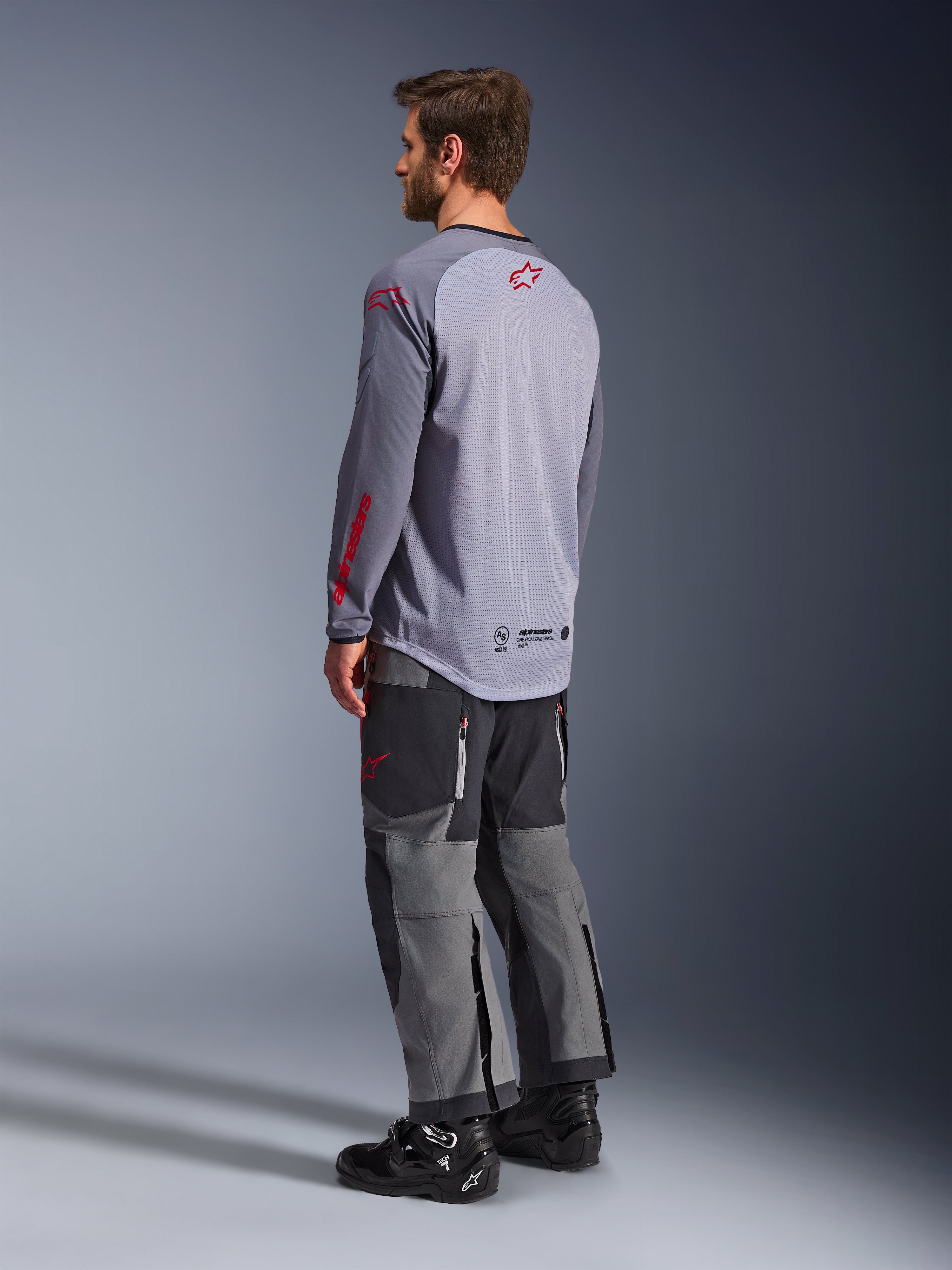 Man som bär Alpinestars Maxdura Dual Jersey, ljusgrå med röda Alpinestars-logotyper, vy bakifrån som visar mesh-torso och långa ärmar, kombinerat med mörkgrå tekniska byxor och svarta motorcykelstövlar