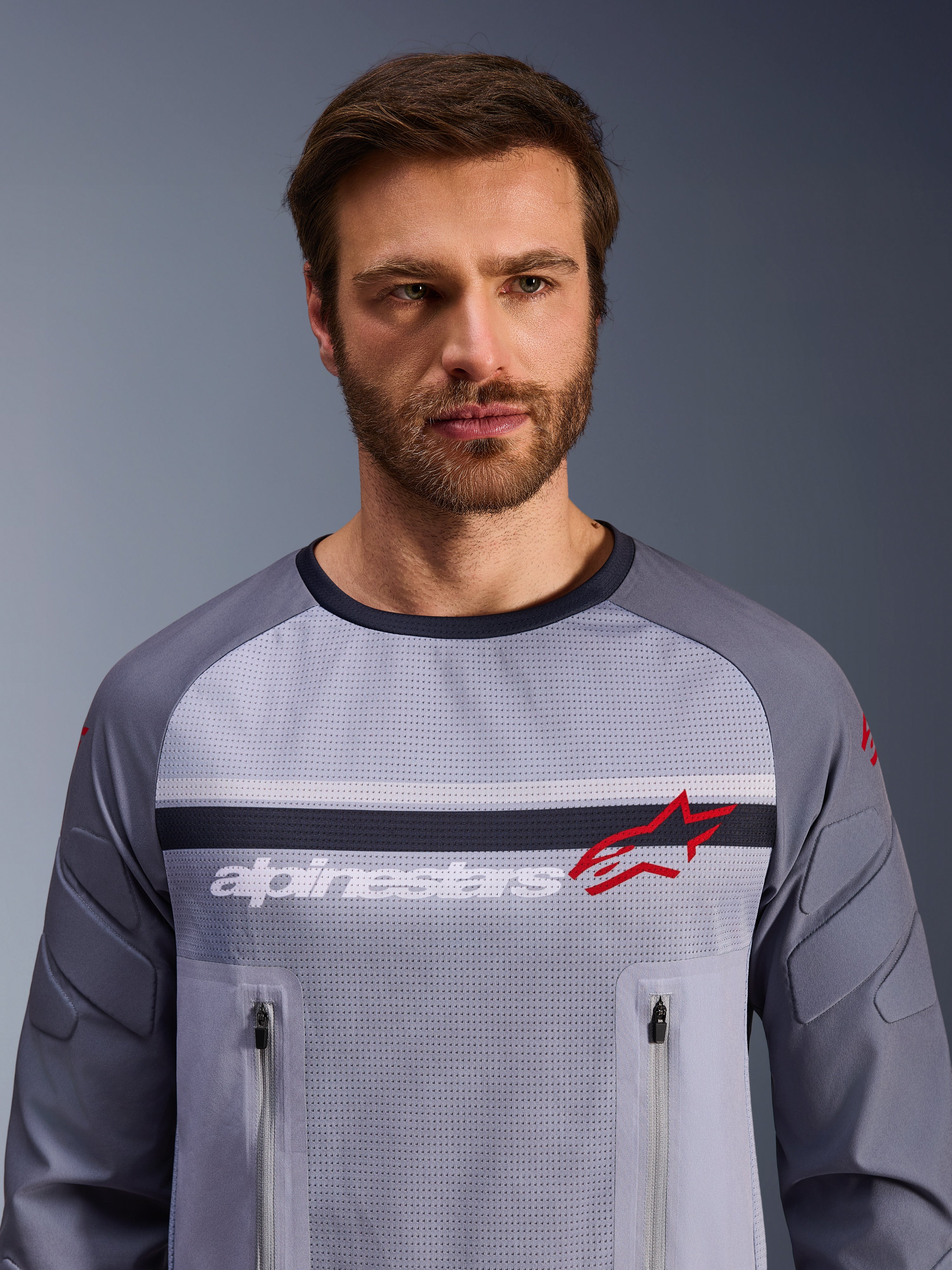 Alpinestars Maxdura Dual Jersey, Mid Gray och Dark Gray, bärs av en manlig modell, med mesh-torso för ventilation, dubbla bröstfickor med dragkedja, vit 'alpinestars'-text och röd stjärnlogotyp, teknisk utrustning för off-road och äventyrskörning