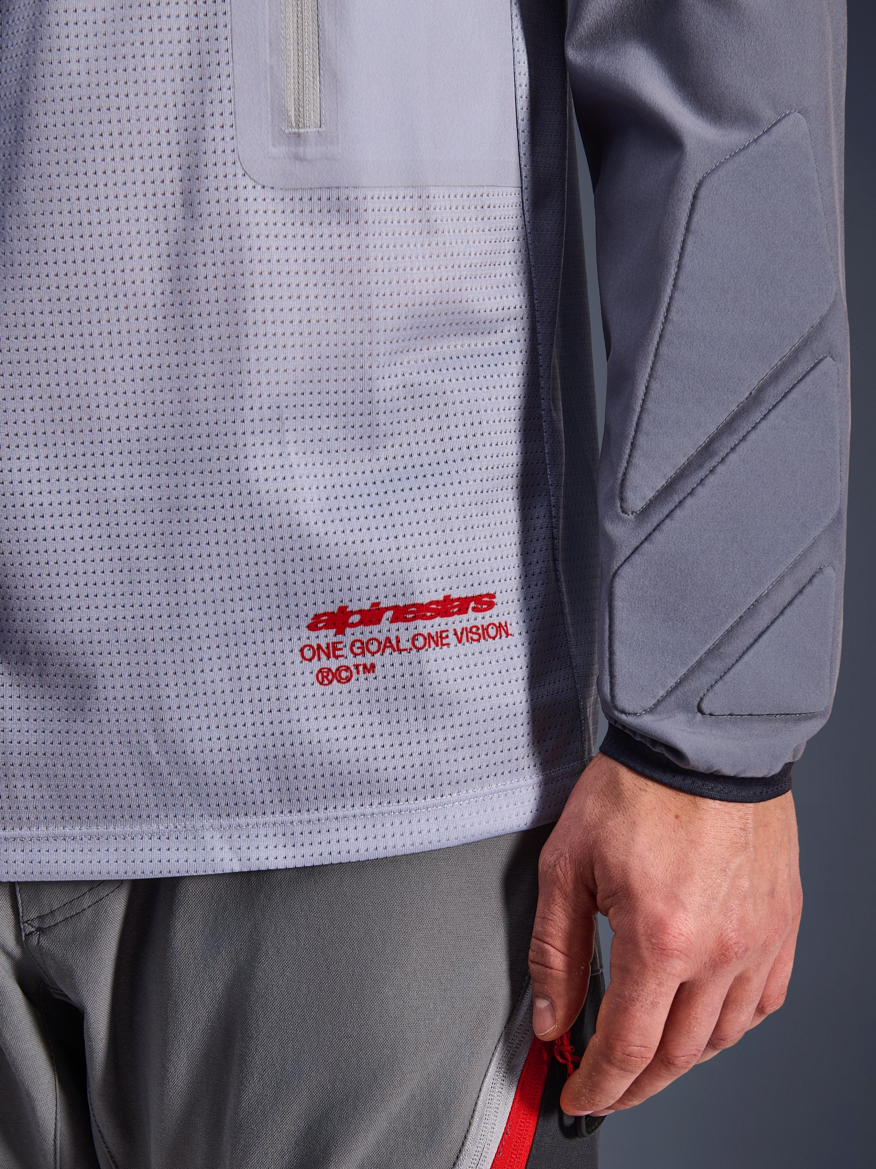 Alpinestars Maxdura Dual Jersey, mellangrå och mörkgrå, närbild som visar ventilerande mesh-bål med röd logotyp, detalj på ficka med dragkedja och integrerad vaddering på ärmen, teknisk motorcykelutrustning för äventyrskörning