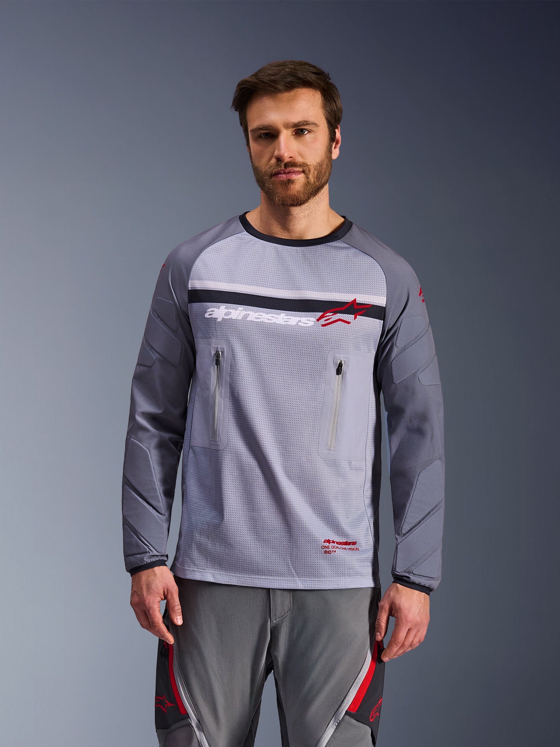 Alpinestars Maxdura Dual Jersey, mellangrå och mörkgrå, bärs av en modell med matchande grå byxor, med bröstfickor med dragkedja, nätkonstruktion på bålen och vadderade nötningsbeständiga ärmar, äventyrsutrustning för terrängkörning med motorcykel