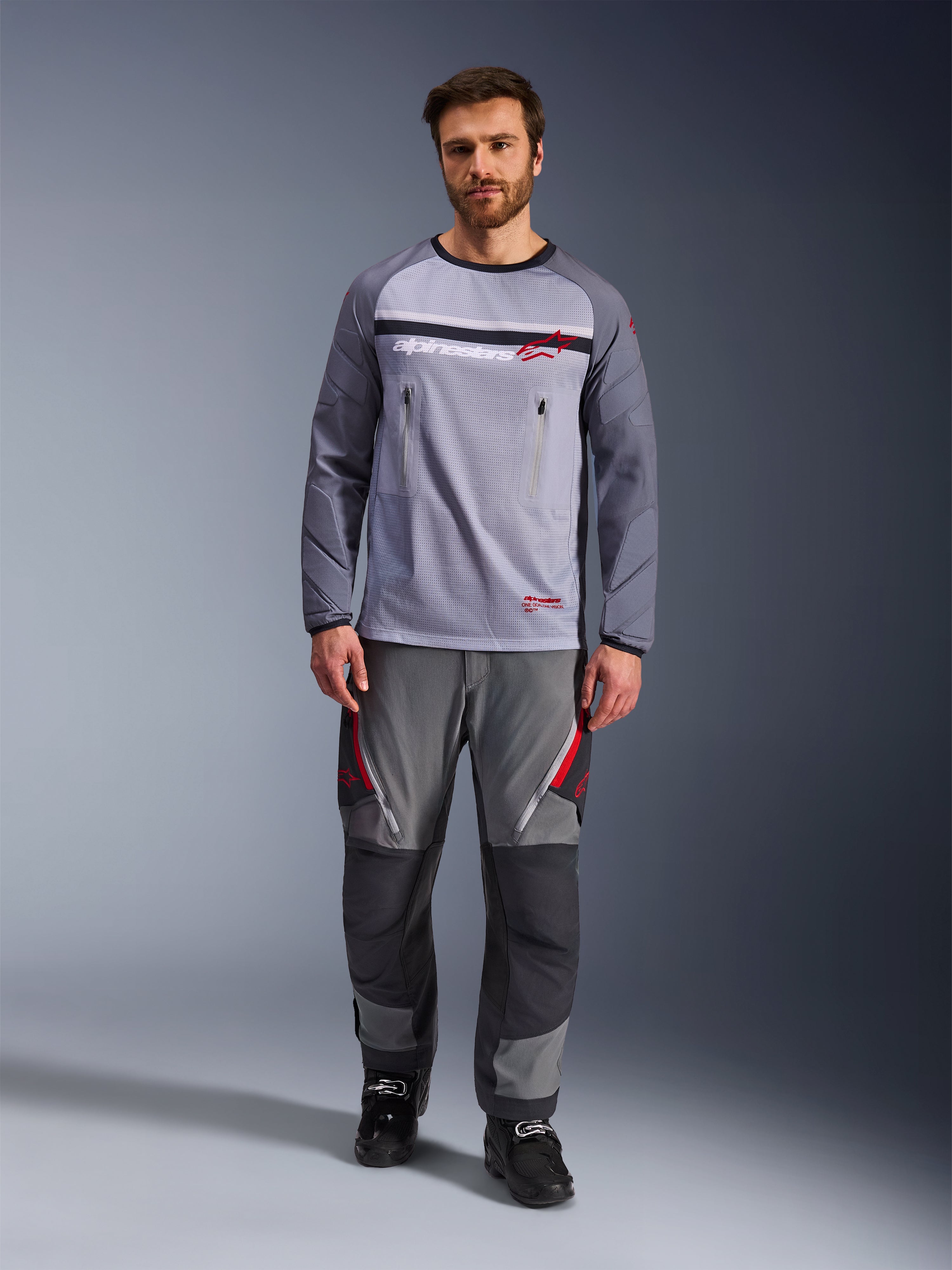 Alpinestars Maxdura Dual Jersey, mellangrå och mörkgrå, äventyrsmotorcykeltröja med torso i mesh och bröstfickor med dragkedja, nötningsbeständiga ärmar med integrerad vaddering, bärs med matchande grå funktionsbyxor med röda detaljer, teknisk terrängkörningsutrustning