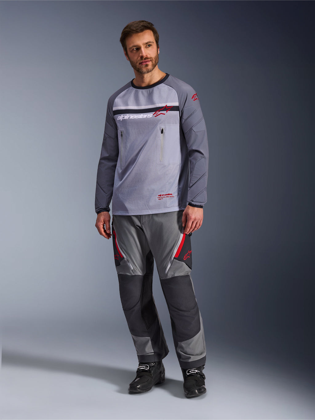 En man som bär Alpinestars Maxdura Dual Jersey i Mid Gray och Dark Gray, stylad med matchande grå och svarta adventurebyxor och svarta stövlar, med en ventilerande mesh-bål, bröstfickor med dragkedja och röda logotypdetaljer.