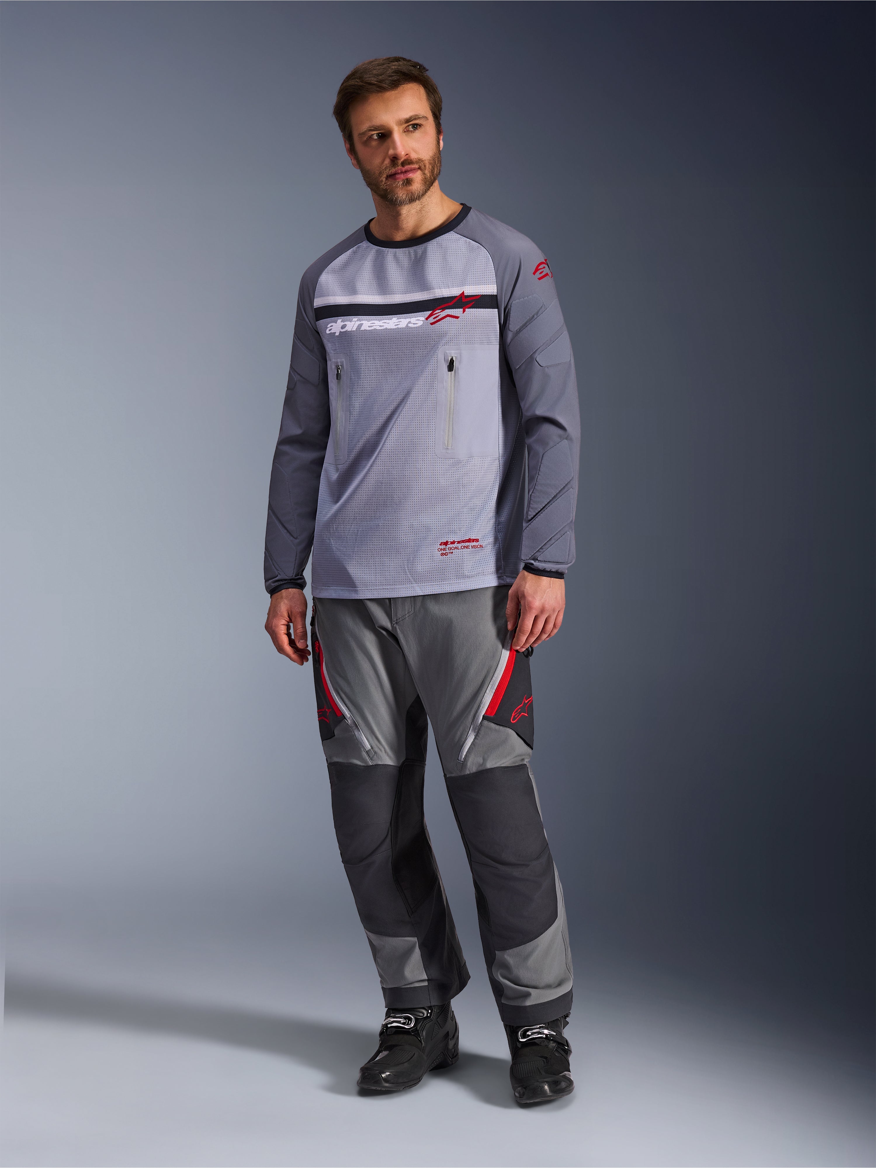 En man som bär Alpinestars Maxdura Dual Jersey i Mid Gray och Dark Gray, stylad med matchande grå och svarta adventurebyxor och svarta stövlar, med en ventilerande mesh-bål, bröstfickor med dragkedja och röda logotypdetaljer.