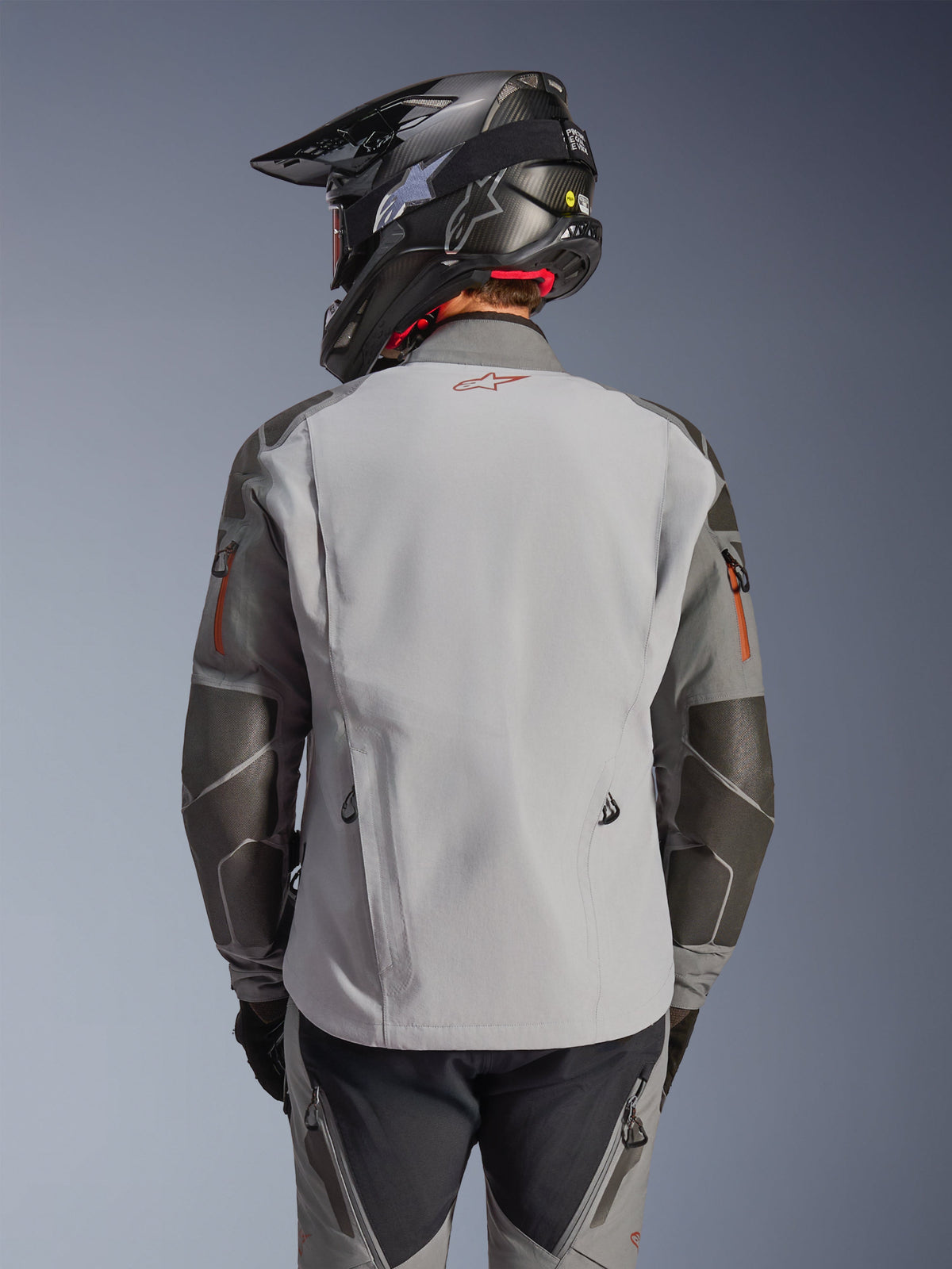 Alpinestars Nazca 3L GORE-TEX PRO-jacka, äventyrsjacka för motorcykel, vy bakifrån, mörkgrå och mellangrå, har Superfabric-förstärkningar på armbågar och axlar, röd logotyp på ryggen, bärs med matchande äventyrsutrustning och en kolfiberhjälm.
