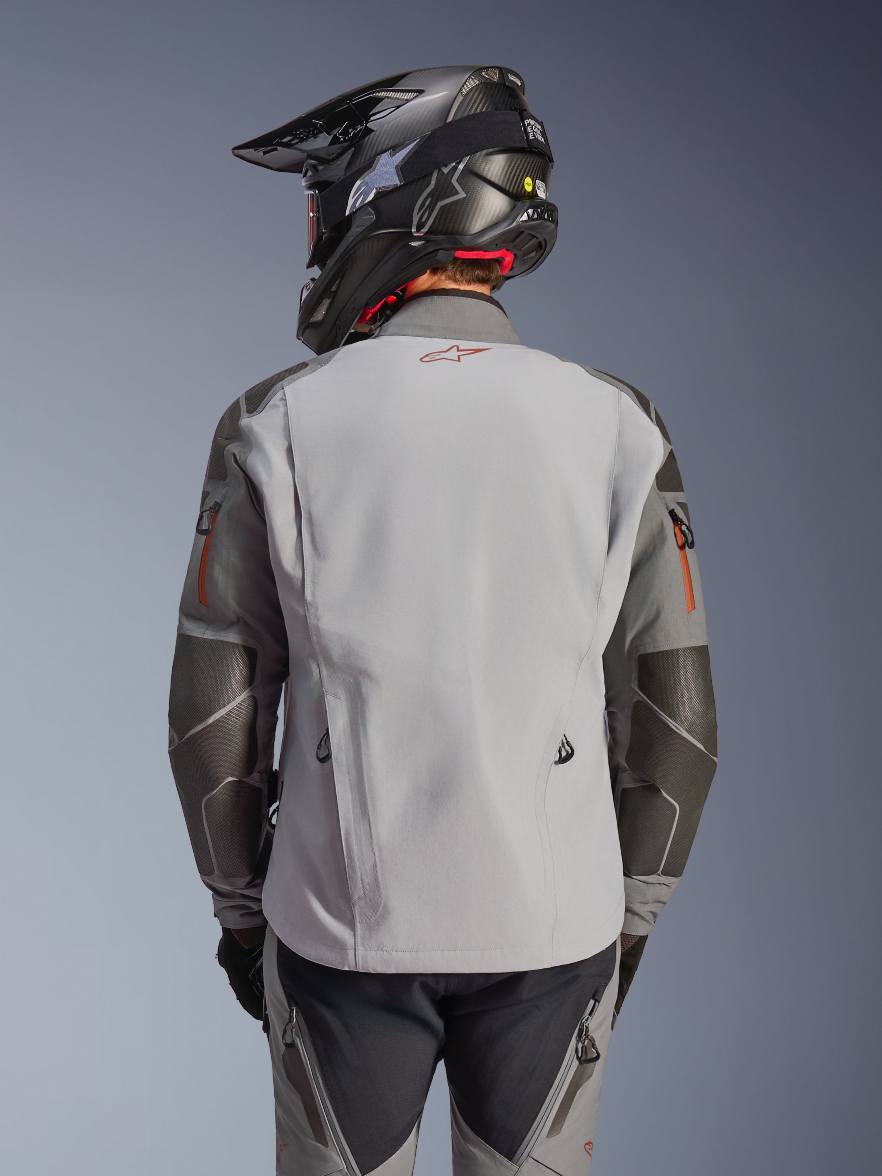 Alpinestars Nazca 3L GORE-TEX PRO-jacka, äventyrsjacka för motorcykel, vy bakifrån, mörkgrå och mellangrå, har Superfabric-förstärkningar på armbågar och axlar, röd logotyp på ryggen, bärs med matchande äventyrsutrustning och en kolfiberhjälm.