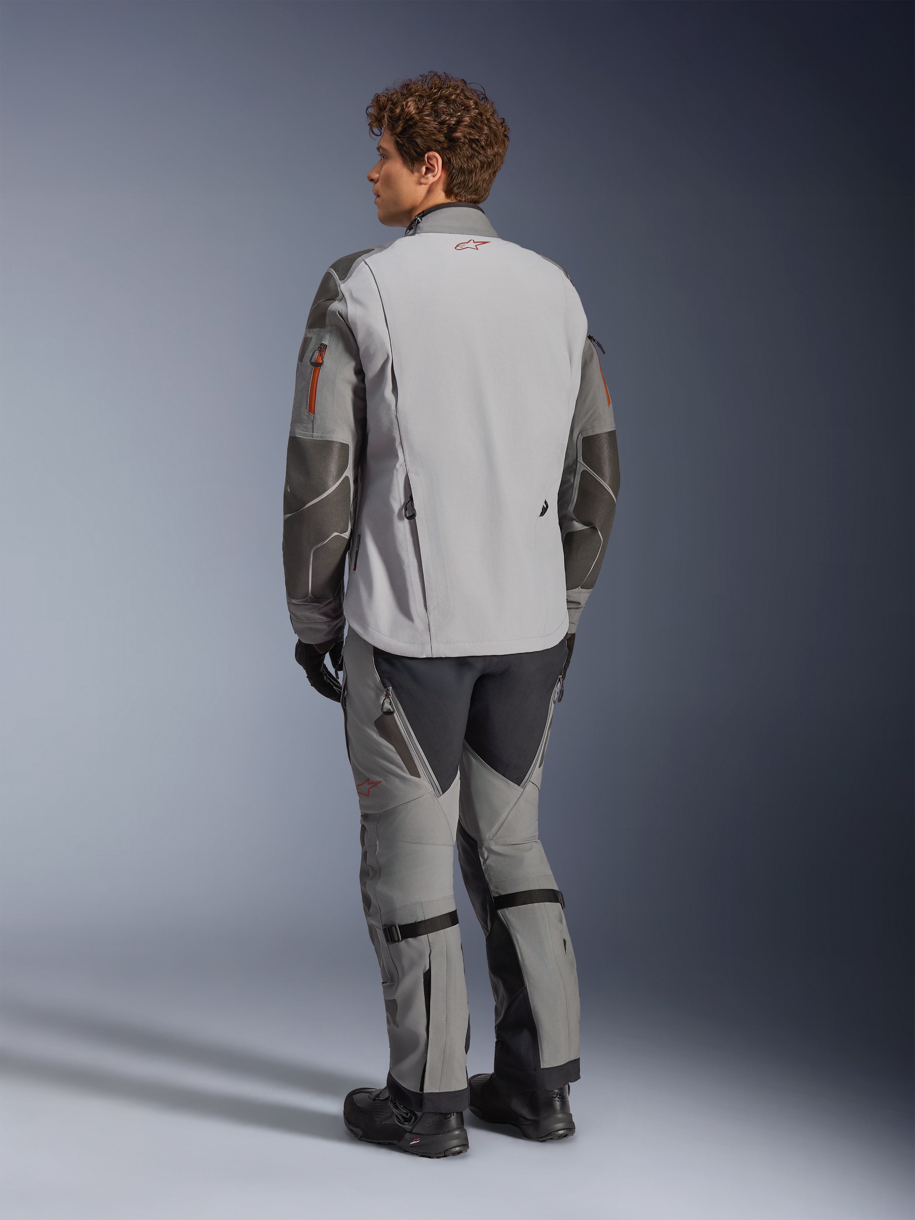 Alpinestars Nazca 3L GORE-TEX PRO-jacka, äventyrsmotorcykeljacka, mörkgrå och mellangrå, vy bakifrån bärs med matchande äventyrsbyxor och svarta stövlar, med mörkgrå Superfabric-förstärkningar på ärmarna, orangea dragkedjedetaljer och en liten röd logotyp på kragen bak.