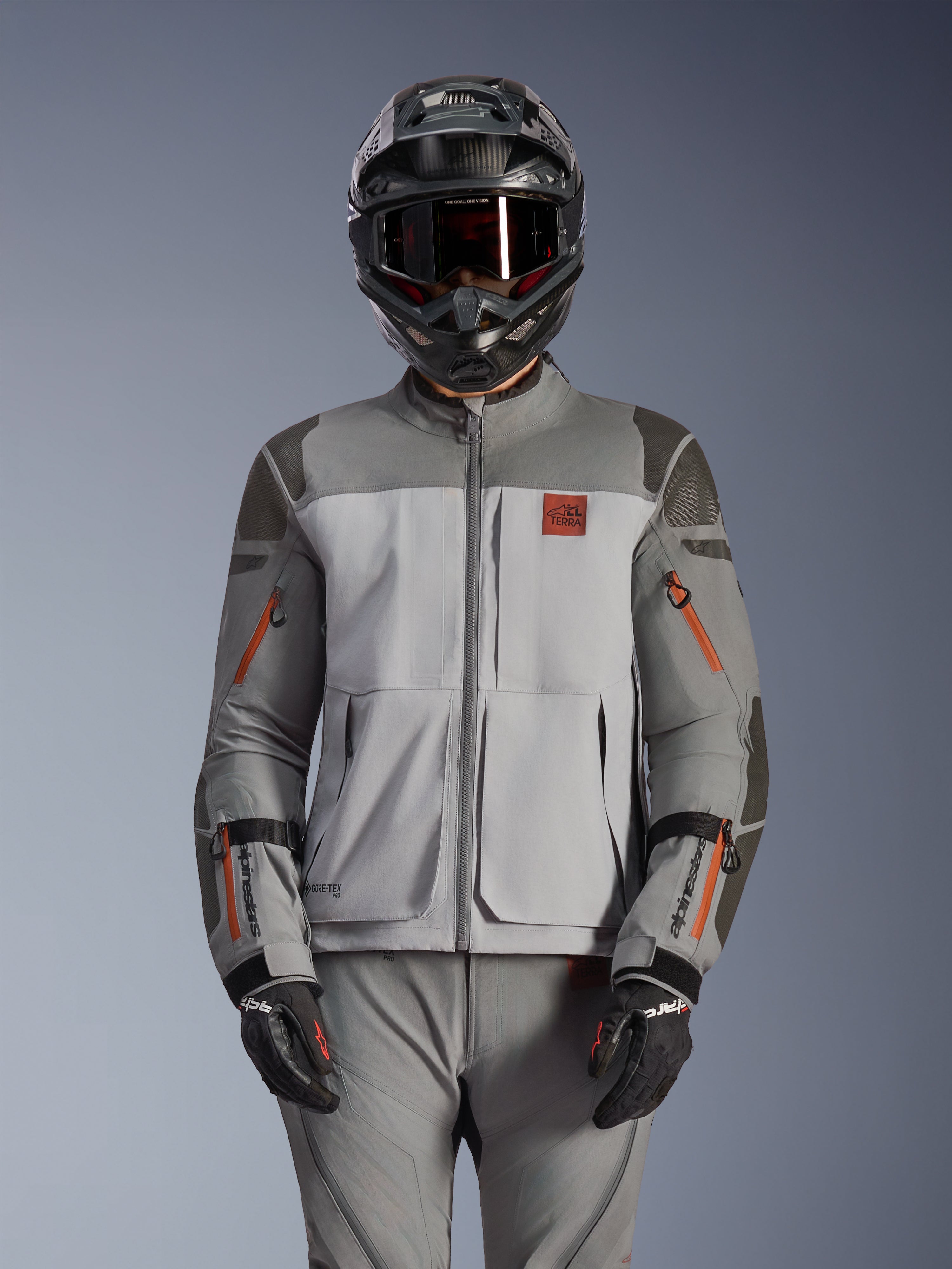 Alpinestars Nazca 3L GORE-TEX PRO-jacka, äventyrsmotorcykeljacka, mörkgrå och mellangrå med orangea dragkedjeaccenter, bärs med matchande byxor och svart hjälm, med hållbar textilkonstruktion och synlig GORE-TEX PRO- och All Terra-branding