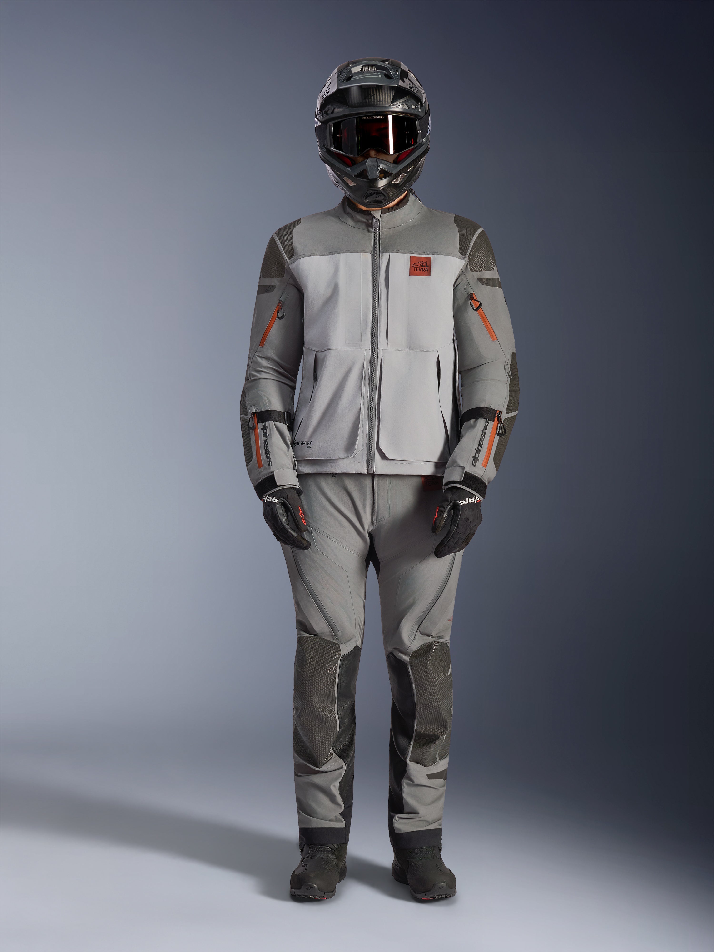 Alpinestars Nazca 3L GORE-TEX PRO-jacka, äventyrsmotorcykeljacka, mörkgrå och mellangrå med orangea detaljer, med Superfabric-förstärkningar och främre ventilationsdragkedjor, bärs med matchande byxor och svart hjälm, teknisk allterrängsutrustning