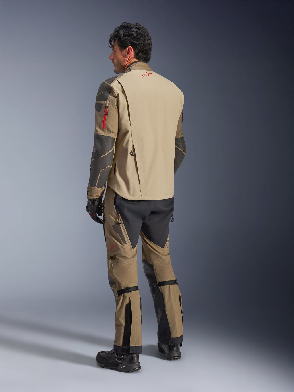 Alpinestars Nazca 3L GORE-TEX PRO-jacka, äventyrsmotorcykeljacka, Walnut Rock Brown, bakifrån-vy av en förare i en komplett äventyrskörningsoutfit med matchande byxor och stövlar, beige och svarta paneler, röd logotyp på övre ryggen, hållbar textilkonstruktion.