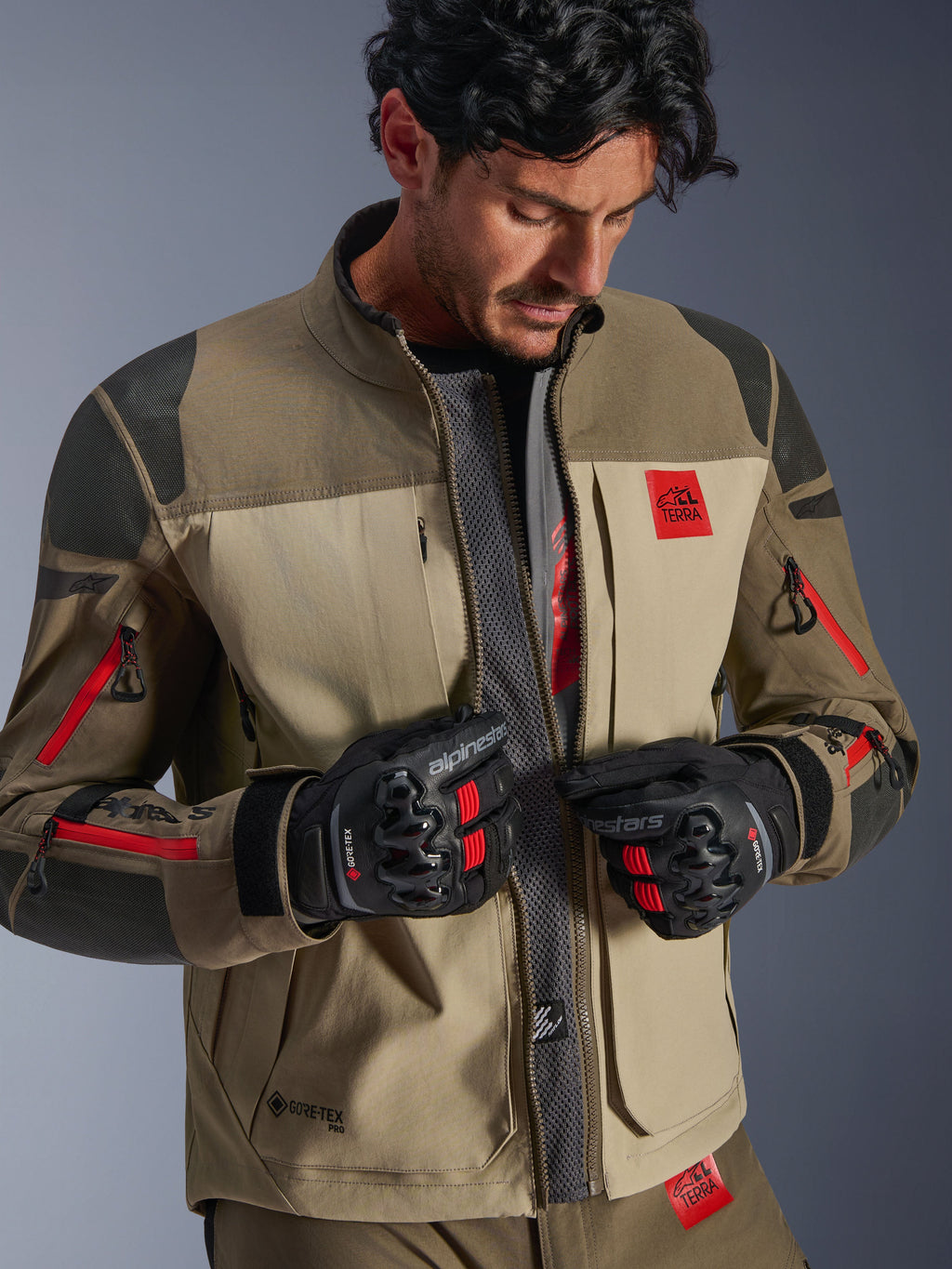 Alpinestars Nazca 3L GORE-TEX PRO-jacka, Walnut Rock Brown, äventyrsmotorcykeljacka med öppen dragkedja som visar central ventilationspanel i mesh, röd All Terra-logotyp, bärs med svarta GORE-TEX-körhandskar, teknisk utrustning för off-road-touring